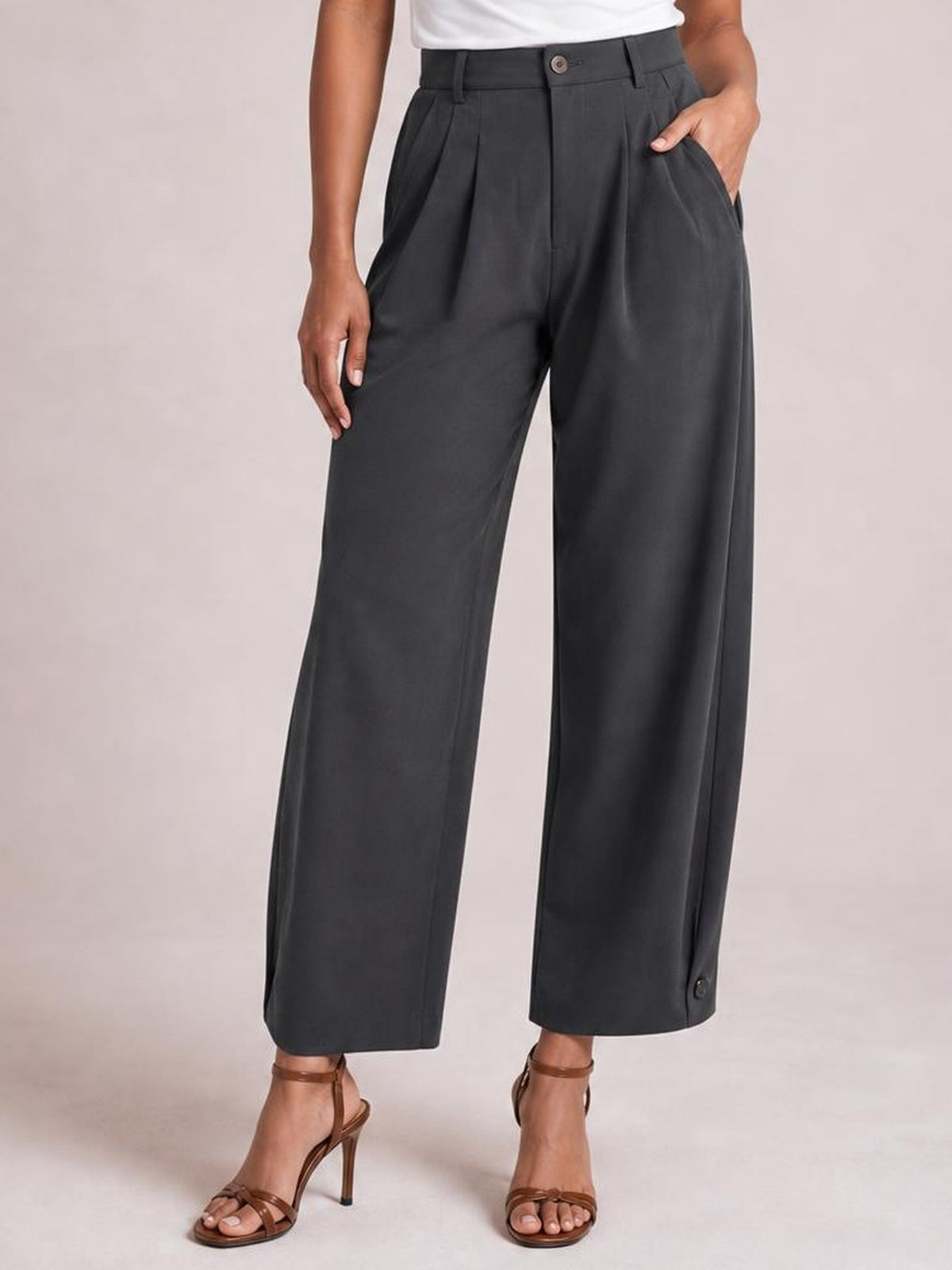 A model wears dark gray high-waisted pleated pants with a button closure, belt loops, slanted pockets, and a relaxed wide-leg ankle-length silhouette.