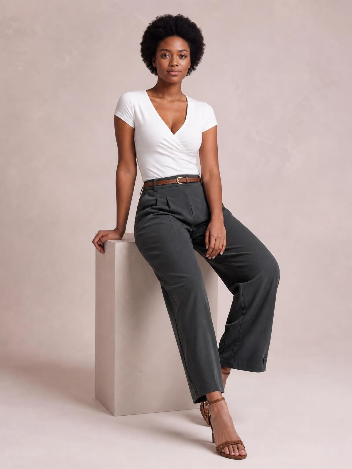 A model sits with one leg crossed, wearing charcoal high-waisted pleated pants with a structured waistband and relaxed wide-leg silhouette.