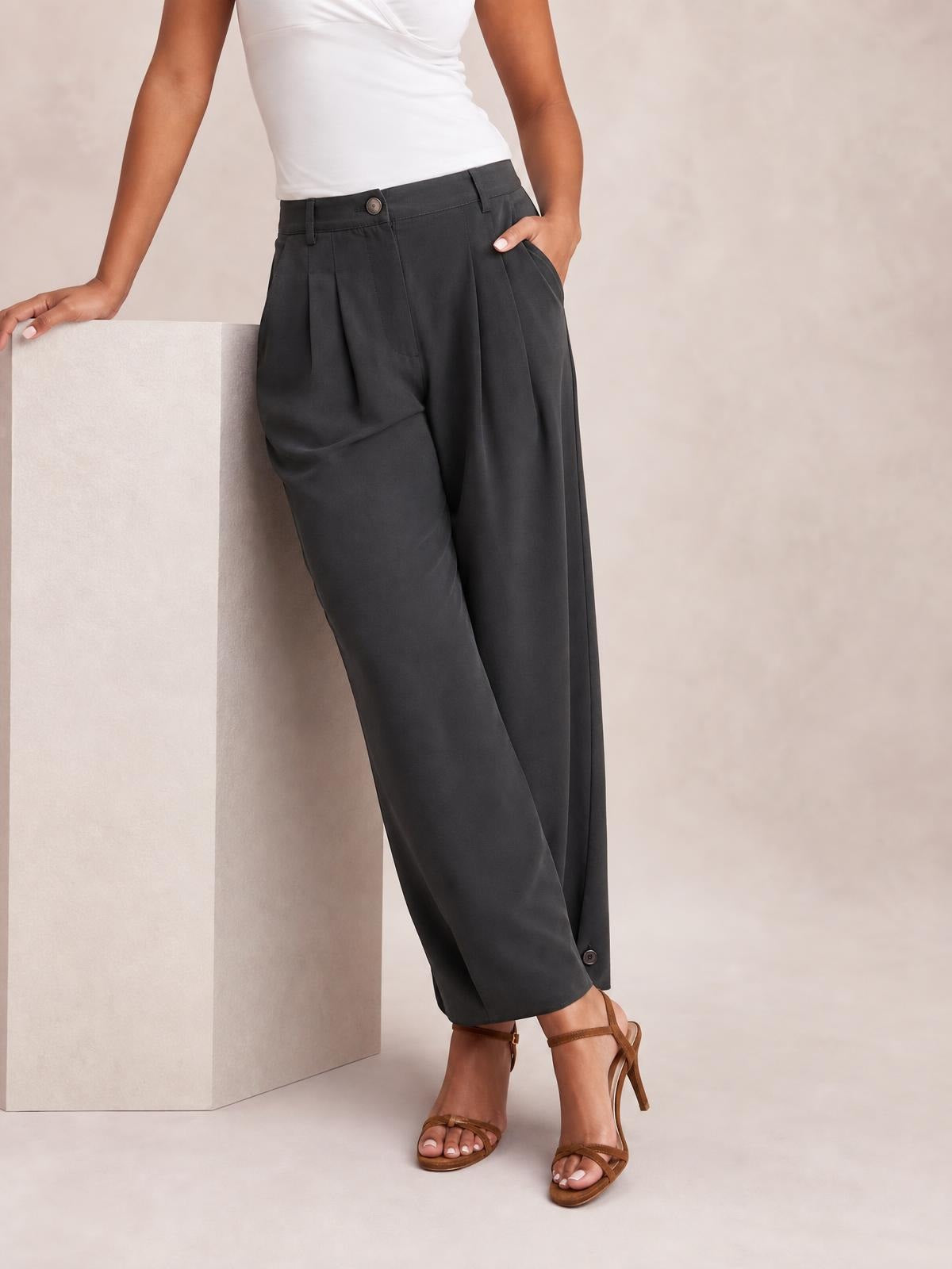A model poses in dark gray high-waisted pleated pants with a relaxed straight-leg silhouette, buttoned waistband, belt loops, and side pockets.