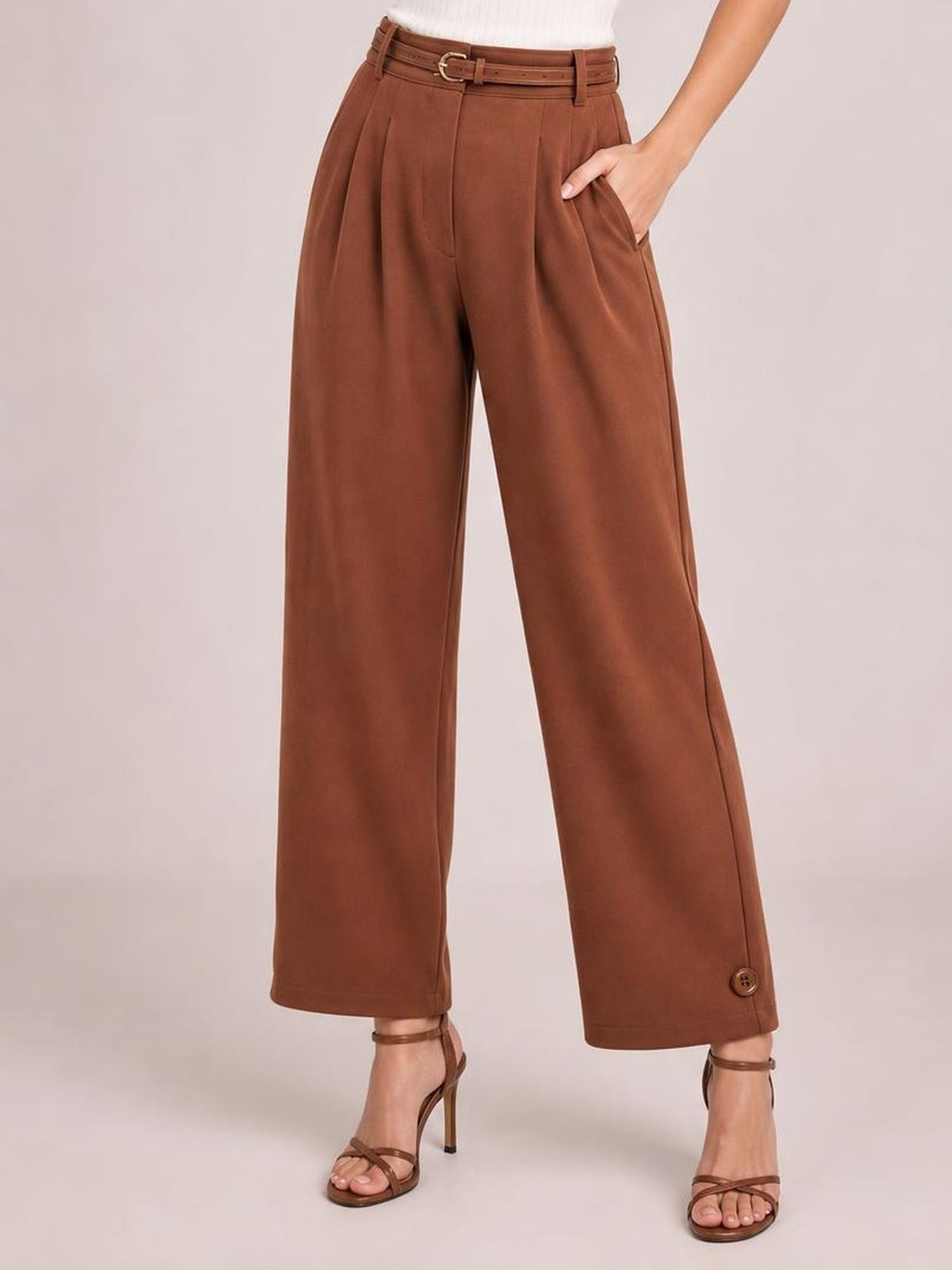 Model wears brown high-waisted pleated pants with a belted waistband, side pockets, wide-leg silhouette, and buttoned ankle hems.