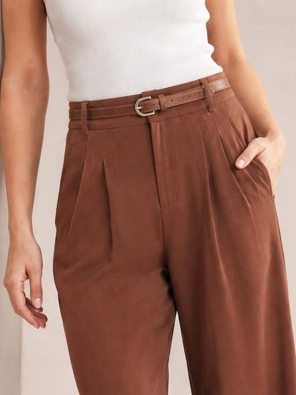 Model wears brown high-waisted pleated pants with a belted waistband, wide-leg silhouette, front pleats, and side pockets, standing with one hand tucked inside a pocket.