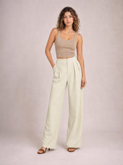 Model poses in cream high-waisted pleated wide-leg pants with front button closure, side pockets, and a relaxed full-length silhouette.