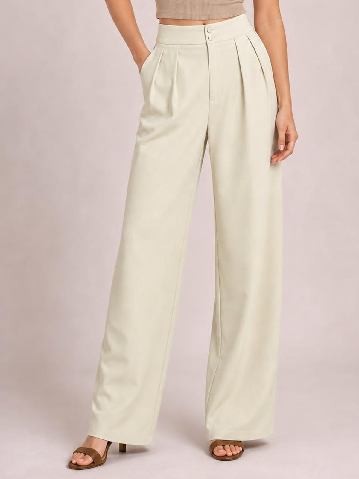 A model wears cream high-waisted pleated wide-leg pants with a buttoned waistband and side pockets, standing with one hand tucked into a pocket.