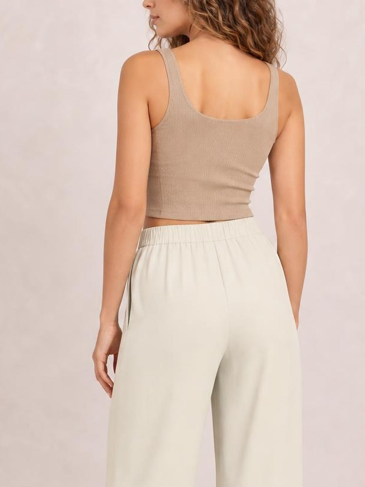 A model shown from behind wears cream high-waisted pleated wide-leg pants with a smooth waistband and side pockets, standing with one hand tucked in a pocket.