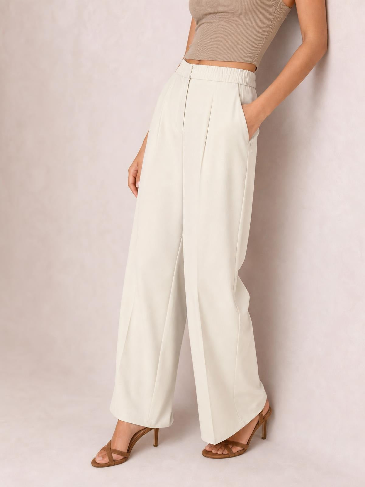 Model wearing high-waisted ivory pleated wide-leg pants with a structured waistband, front button closure, side pockets, and a relaxed flowing silhouette.