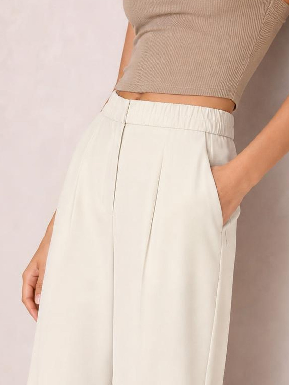 A model poses with one hand in the pocket of cream high-waisted pleated wide-leg pants, showing the relaxed silhouette and structured front seams.