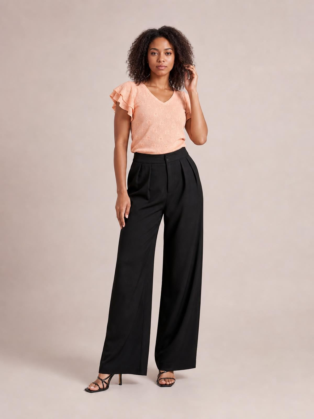 Model poses in black high-waisted pleated wide-leg pants with a buttoned waistband, relaxed flowing silhouette, and visible side pockets.
