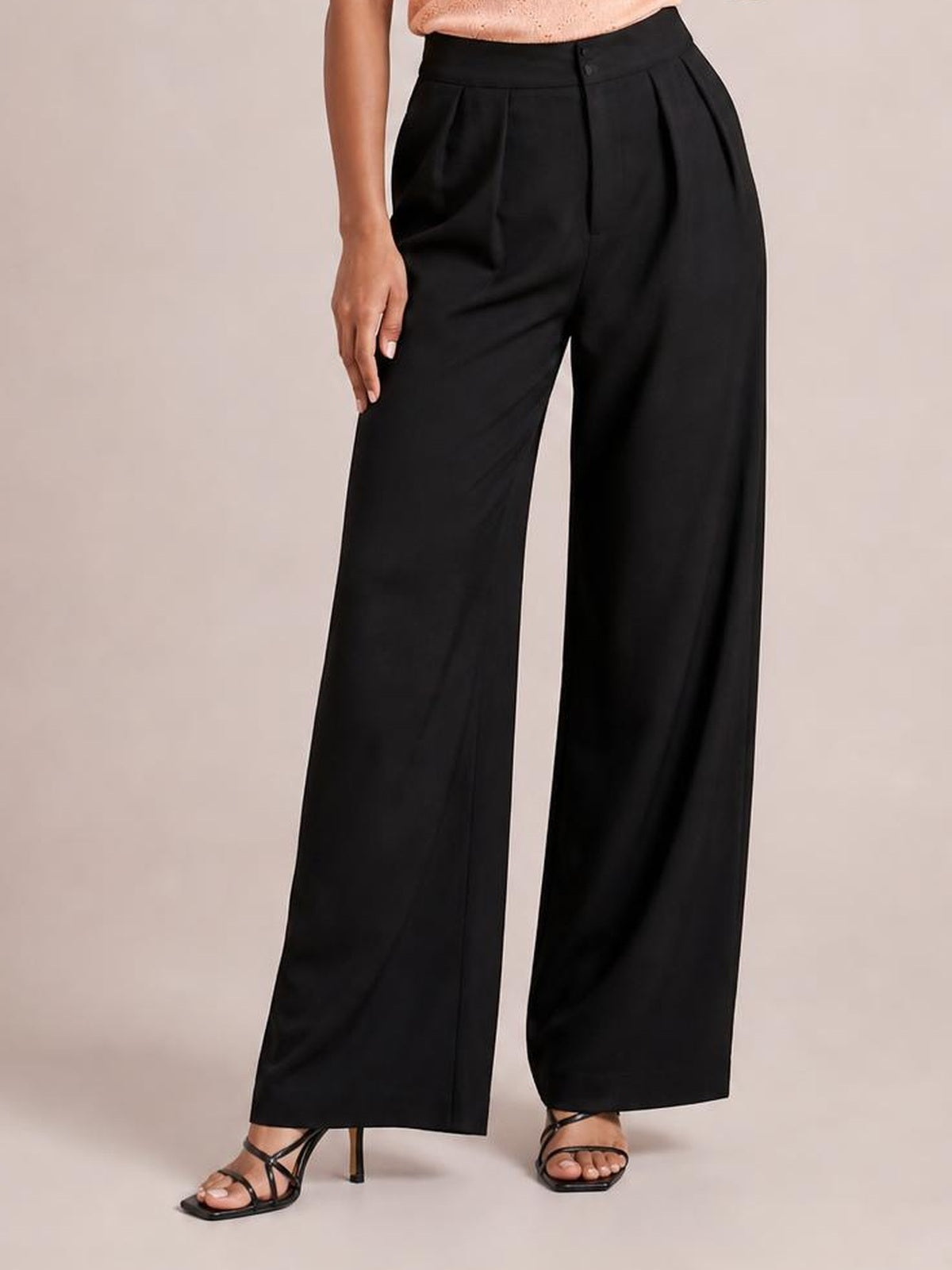 Model wearing black high-waisted pleated wide-leg pants with a button closure and relaxed floor-length silhouette, posed with one hand resting on the thigh.