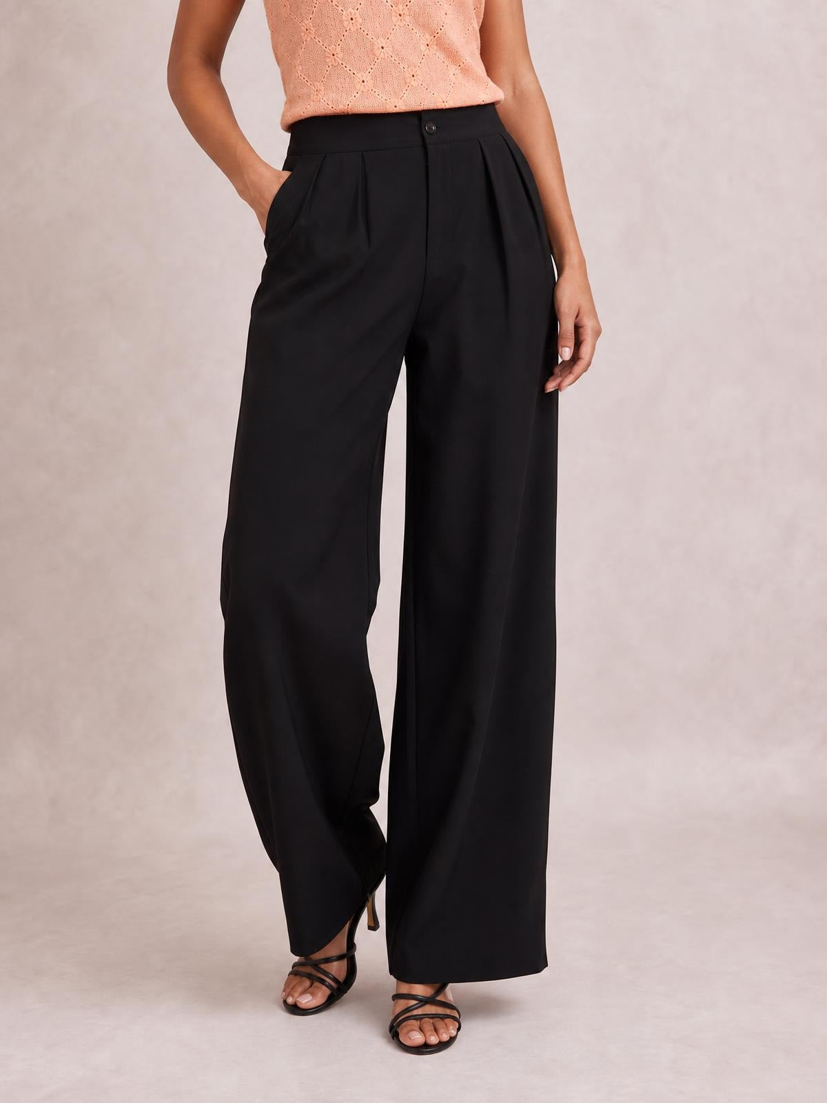 A model poses in black high-waisted pleated wide-leg pants with a buttoned waistband, side pockets, and a flowing full-length silhouette.