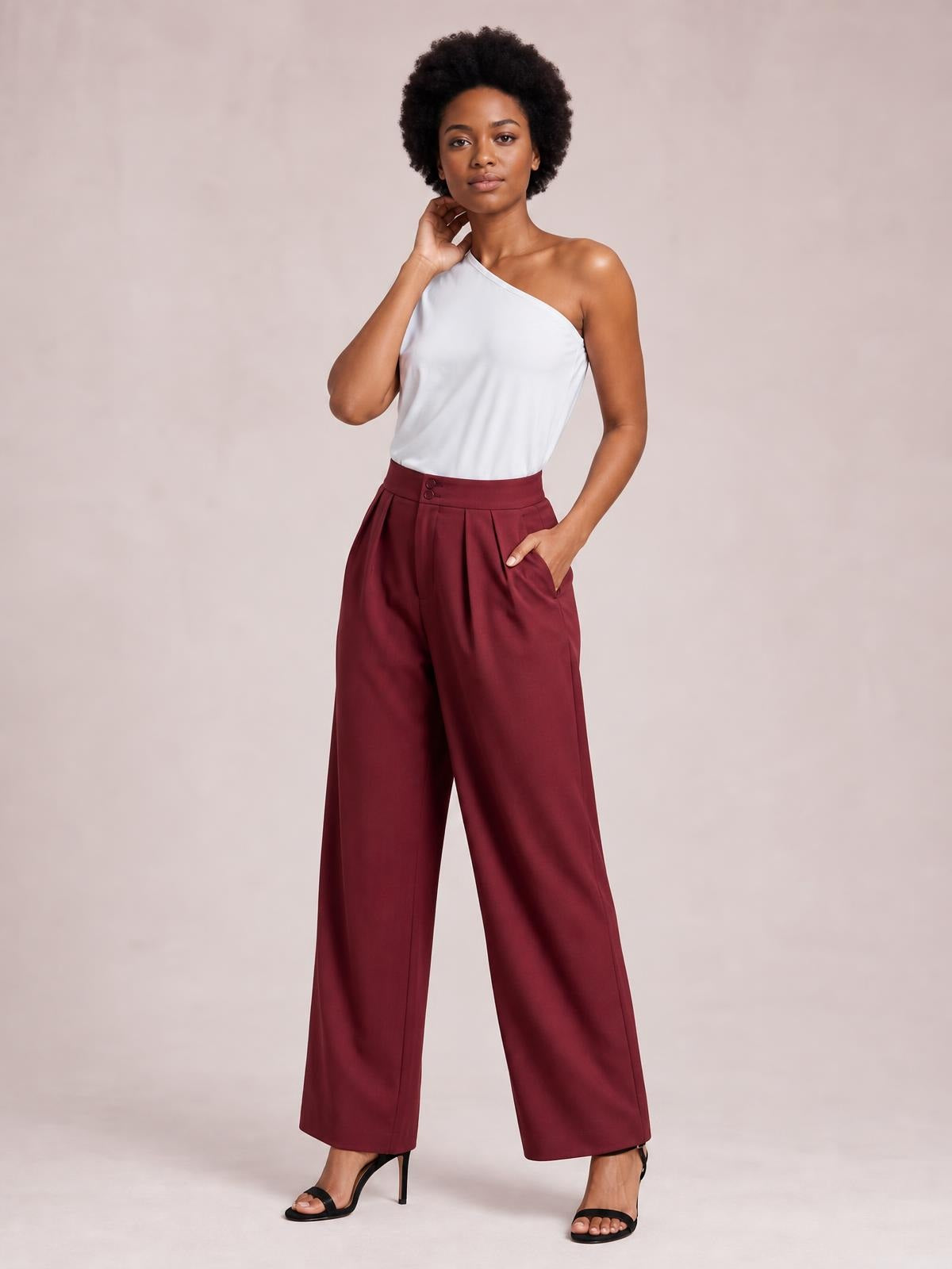 Model wearing burgundy high-waisted pleated wide-leg pants with buttoned waistband and side pockets, posing with one hand tucked into a pocket.