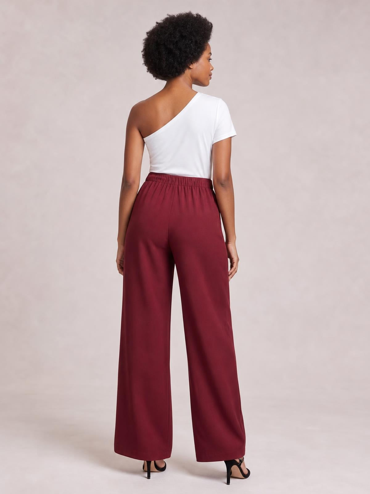 A model stands facing away in burgundy high-waisted pleated wide-leg pants with a gathered waistband and fluid, full-length silhouette.