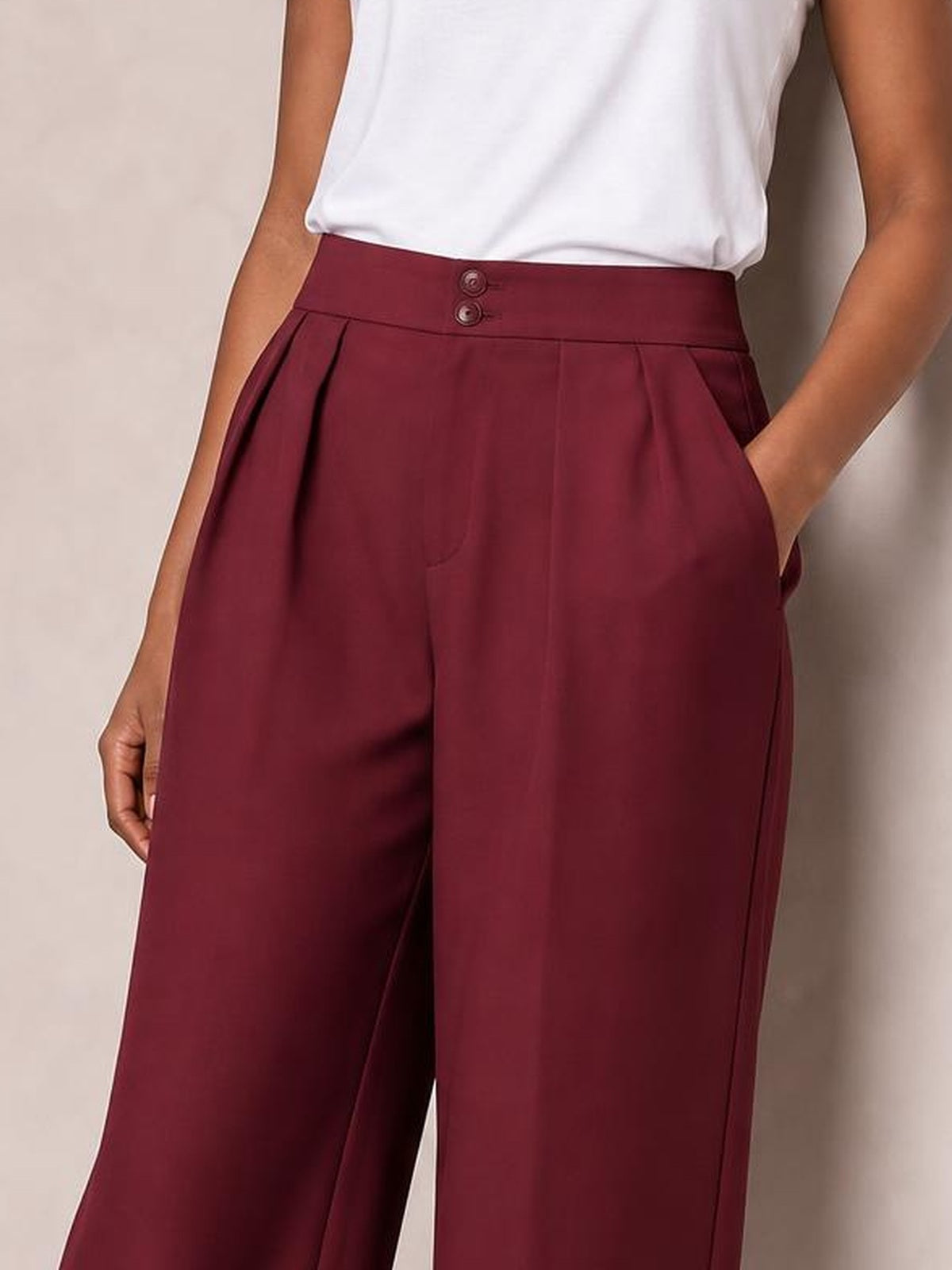 A model wears burgundy high-waisted pleated wide-leg pants with a double-button waistband and side pockets, standing with both hands tucked into the pockets.
