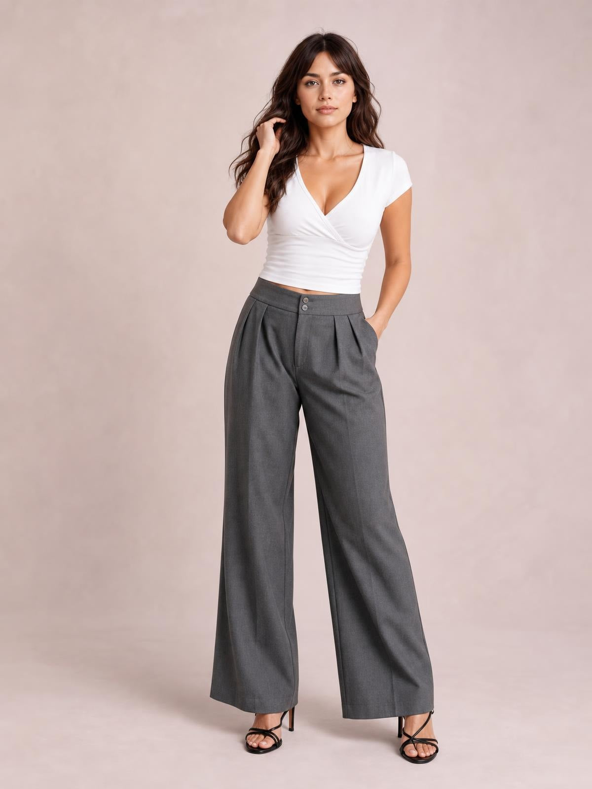 Model wearing gray high-waisted pleated wide-leg pants with front buttons and side pockets, standing with one hand in a pocket.