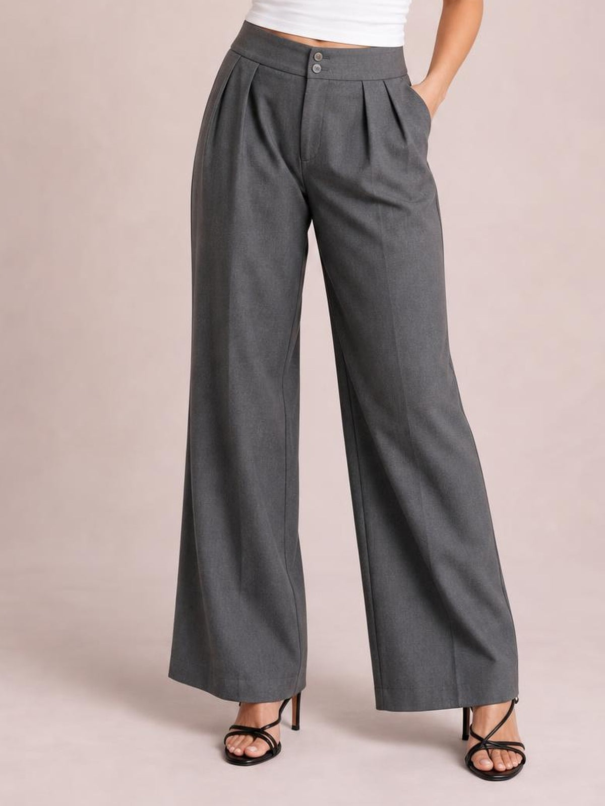 Model wearing gray high-waisted pleated wide-leg pants with a buttoned waistband, side pockets, and a relaxed flowing silhouette.