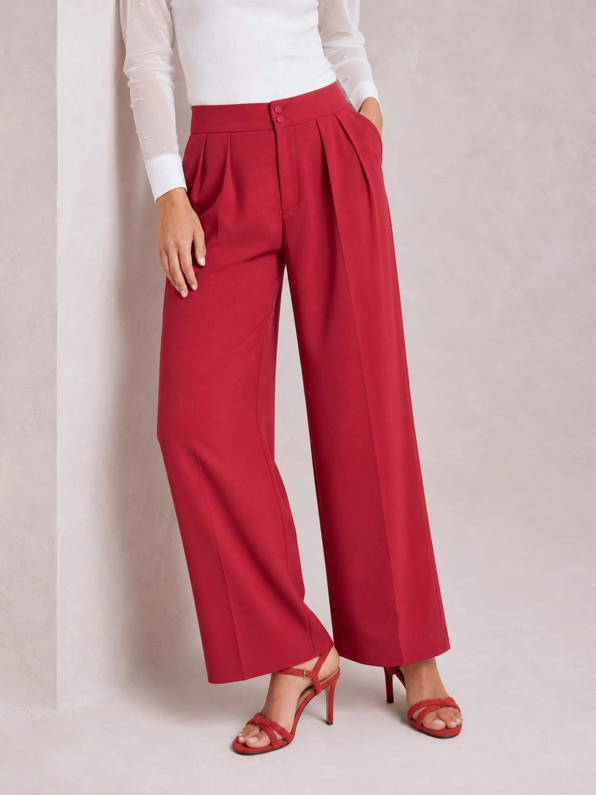 A model poses in red high-waisted pleated wide-leg pants with a buttoned front, side pockets, and a flowing full-length silhouette.