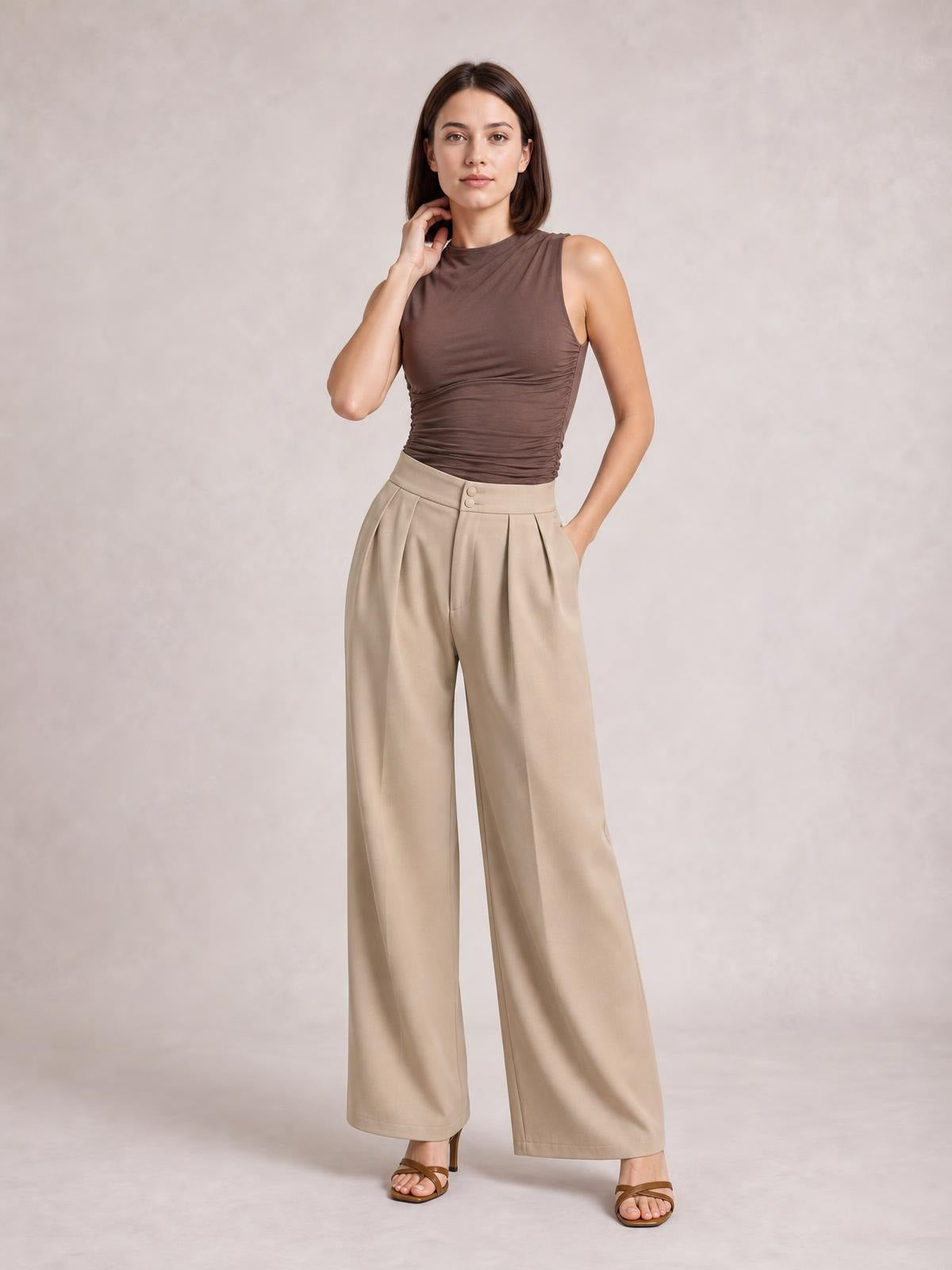 A model poses in beige high-waisted pleated wide-leg pants with a buttoned waistband and side pockets, standing with one hand tucked into a pocket.