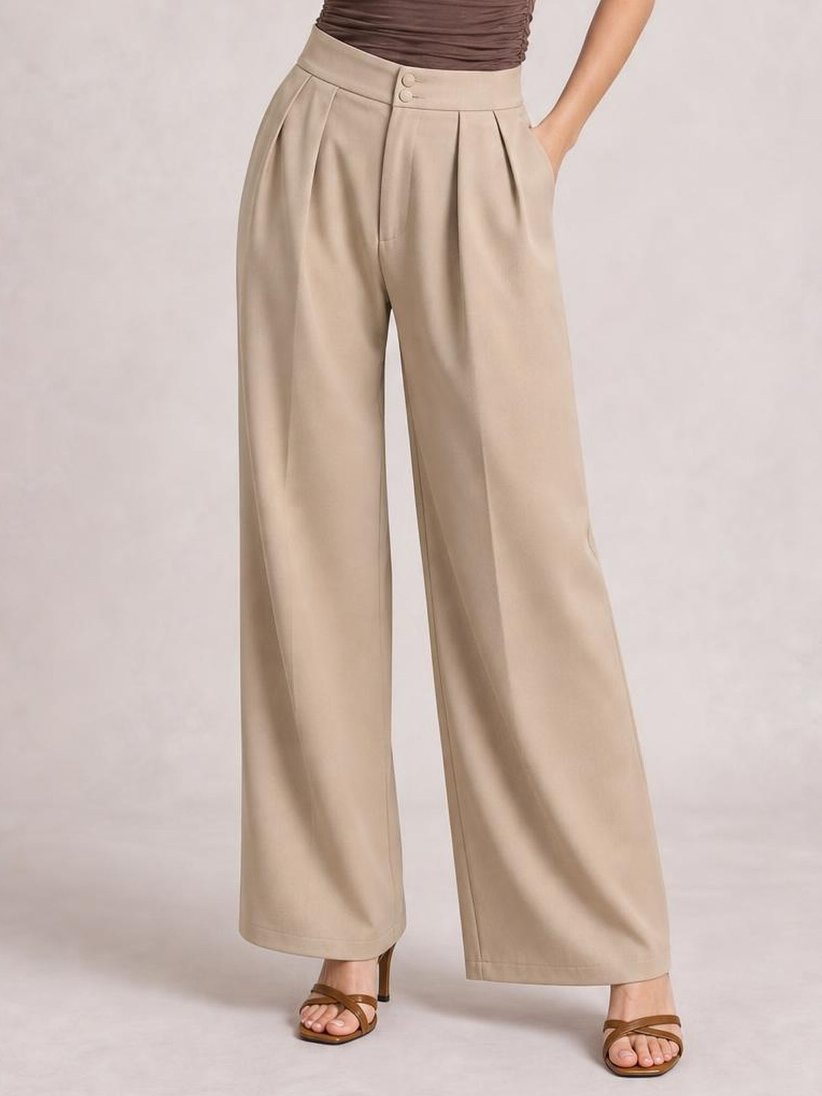 A model wears beige high-waisted pleated wide-leg pants with a buttoned waistband and side pockets, posing with one hand tucked into a pocket.