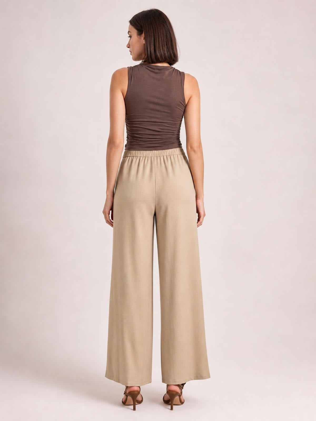 Model shown from the back wears beige high-waisted wide-leg pants with a flowing silhouette and gathered waistband, paired with a fitted sleeveless top.