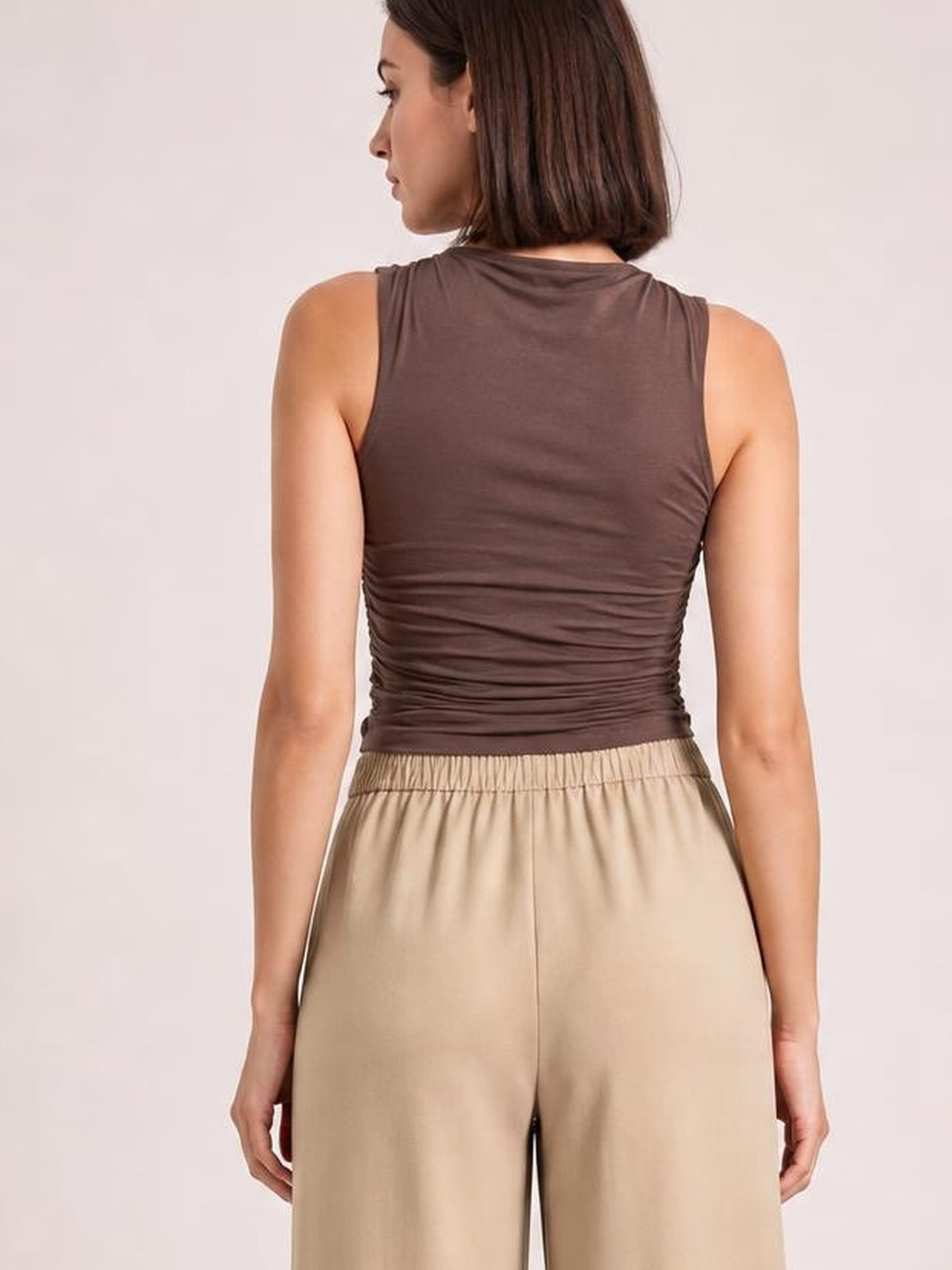 A model stands in a rear view wearing beige high-waisted pleated wide-leg pants with a relaxed silhouette and smooth, flowing construction.