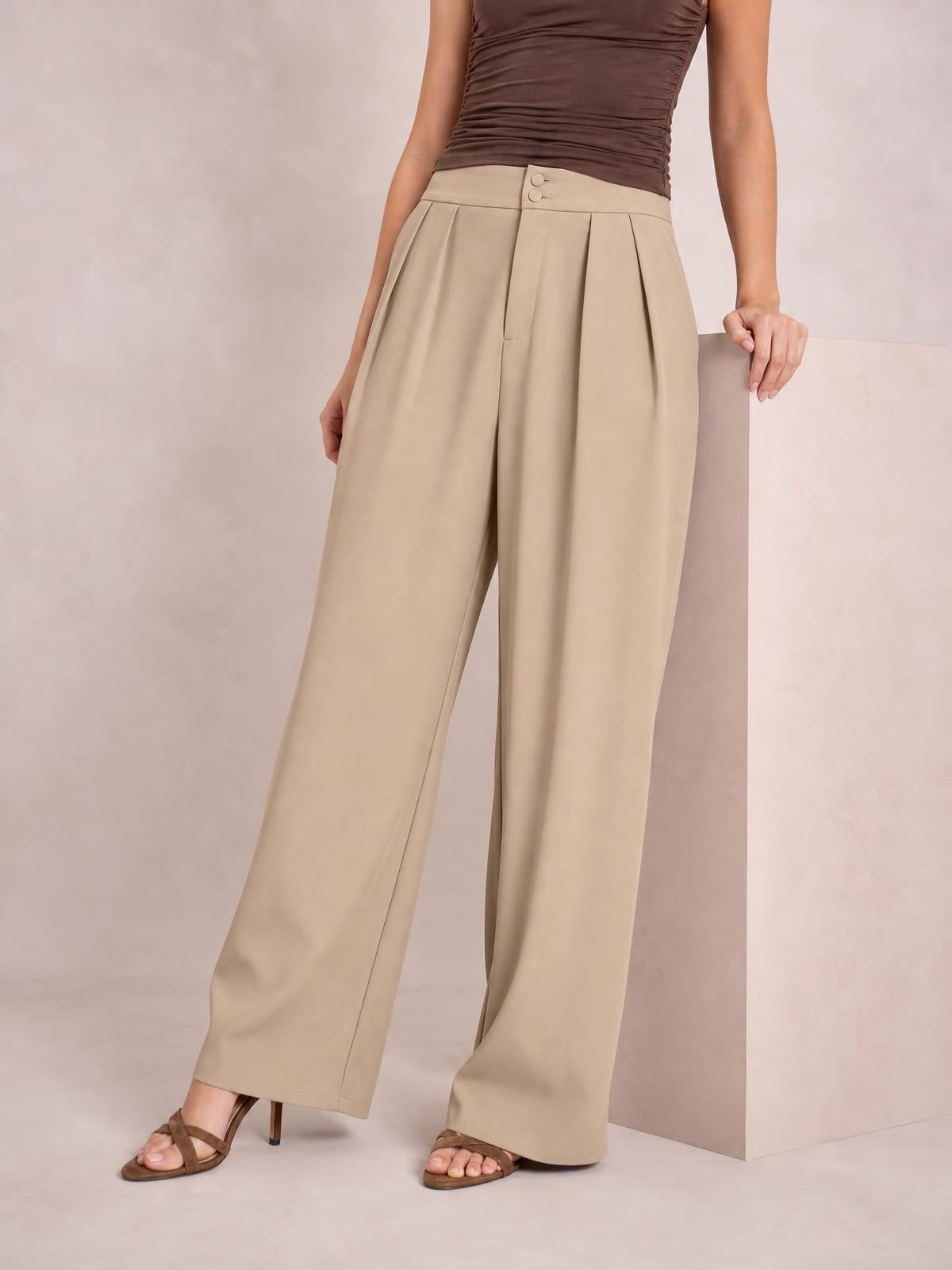 A model wears beige high-waisted pleated wide-leg pants with a buttoned waistband, slant pockets, and a fluid full-length silhouette.