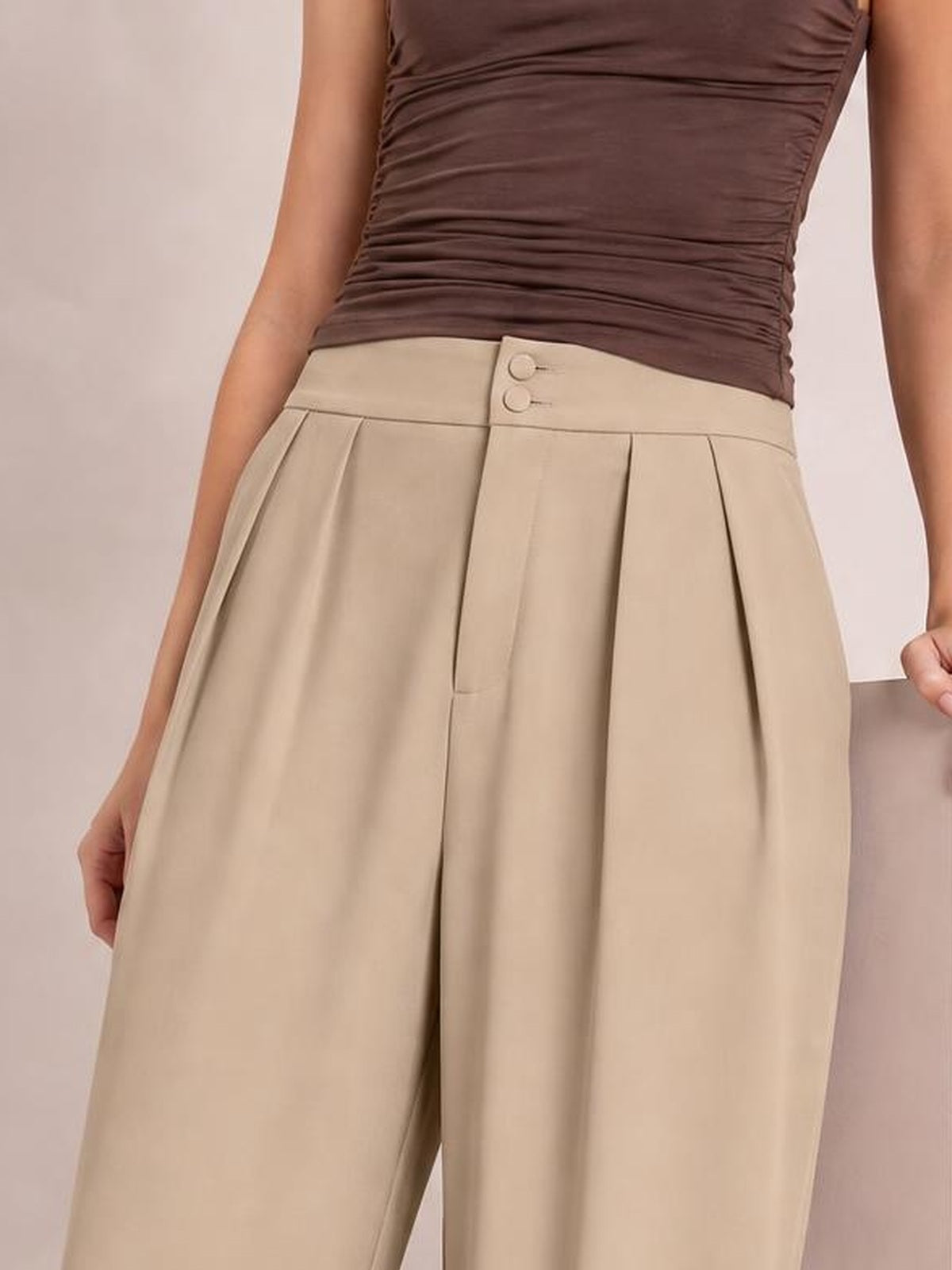 A cropped model wears beige high-waisted pleated wide-leg pants with a buttoned waistband, front pleats, and side pockets, standing with hands at her sides.