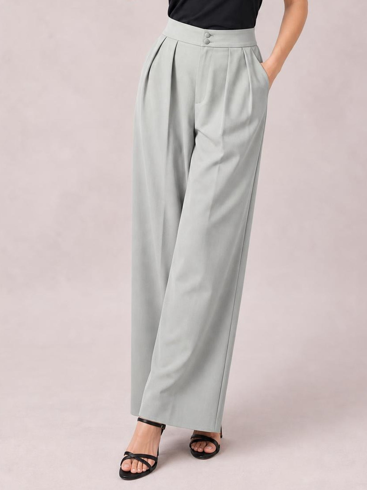 Model poses with one hand in the pocket of light gray high-waisted pleated wide-leg pants, featuring a buttoned waistband and flowing full-length silhouette.