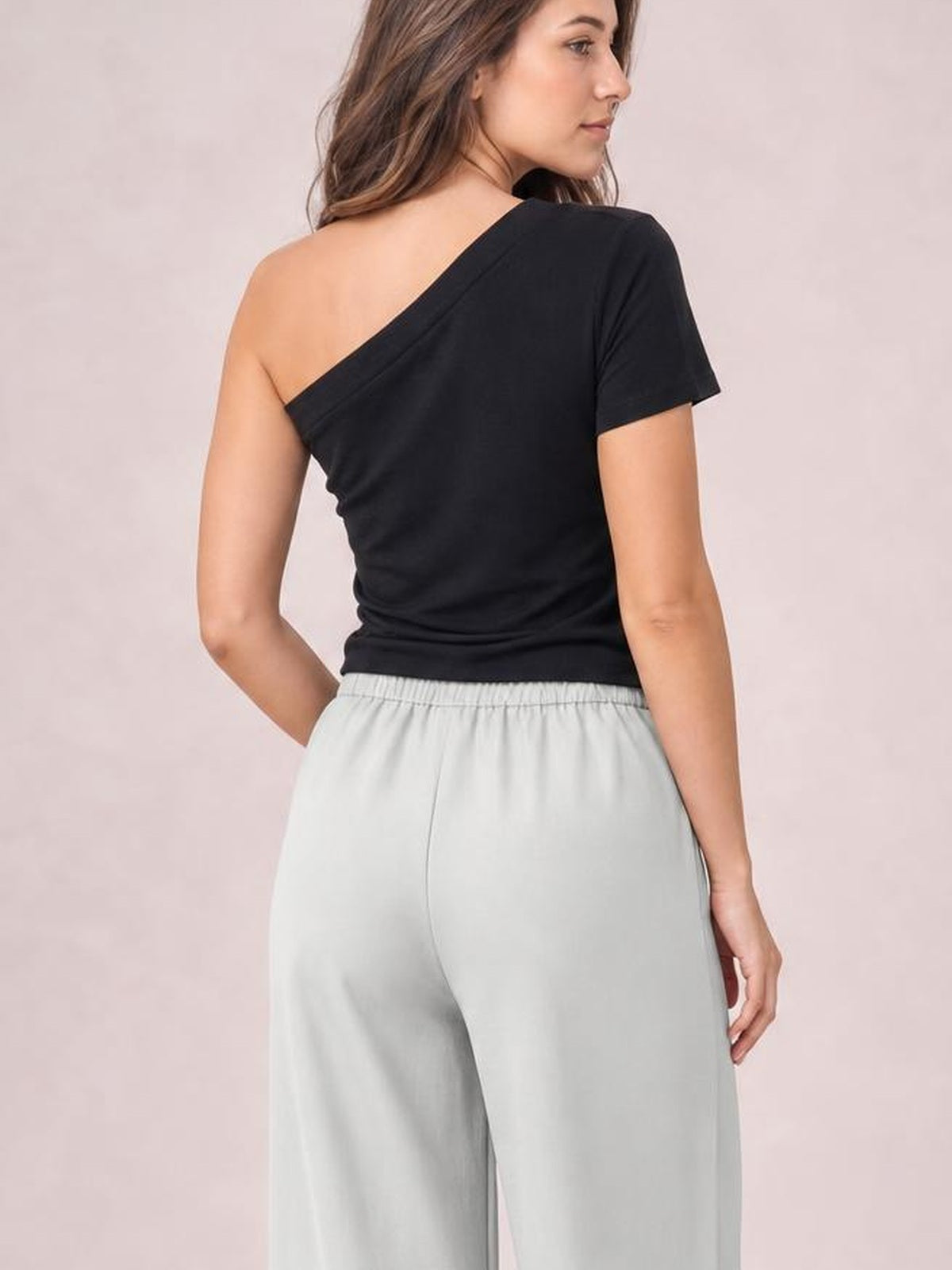 A model poses from behind in light gray high-waisted wide-leg pants with a gathered waistband, relaxed flowing silhouette, and one hand in a pocket.