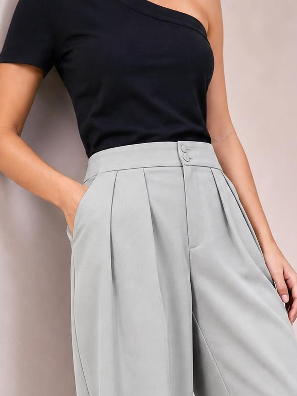 A model wears light gray high-waisted pleated wide-leg pants with a structured waistband, button closure, and side pockets, posing with one hand tucked in.