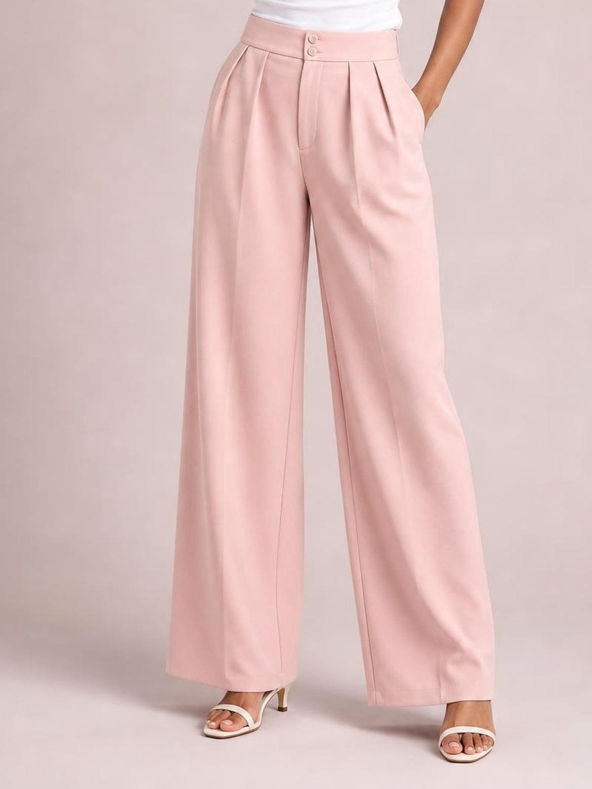 A model poses with one hand in a pocket, wearing pink high-waisted pleated wide-leg pants with a buttoned waistband and flowing full-length silhouette.