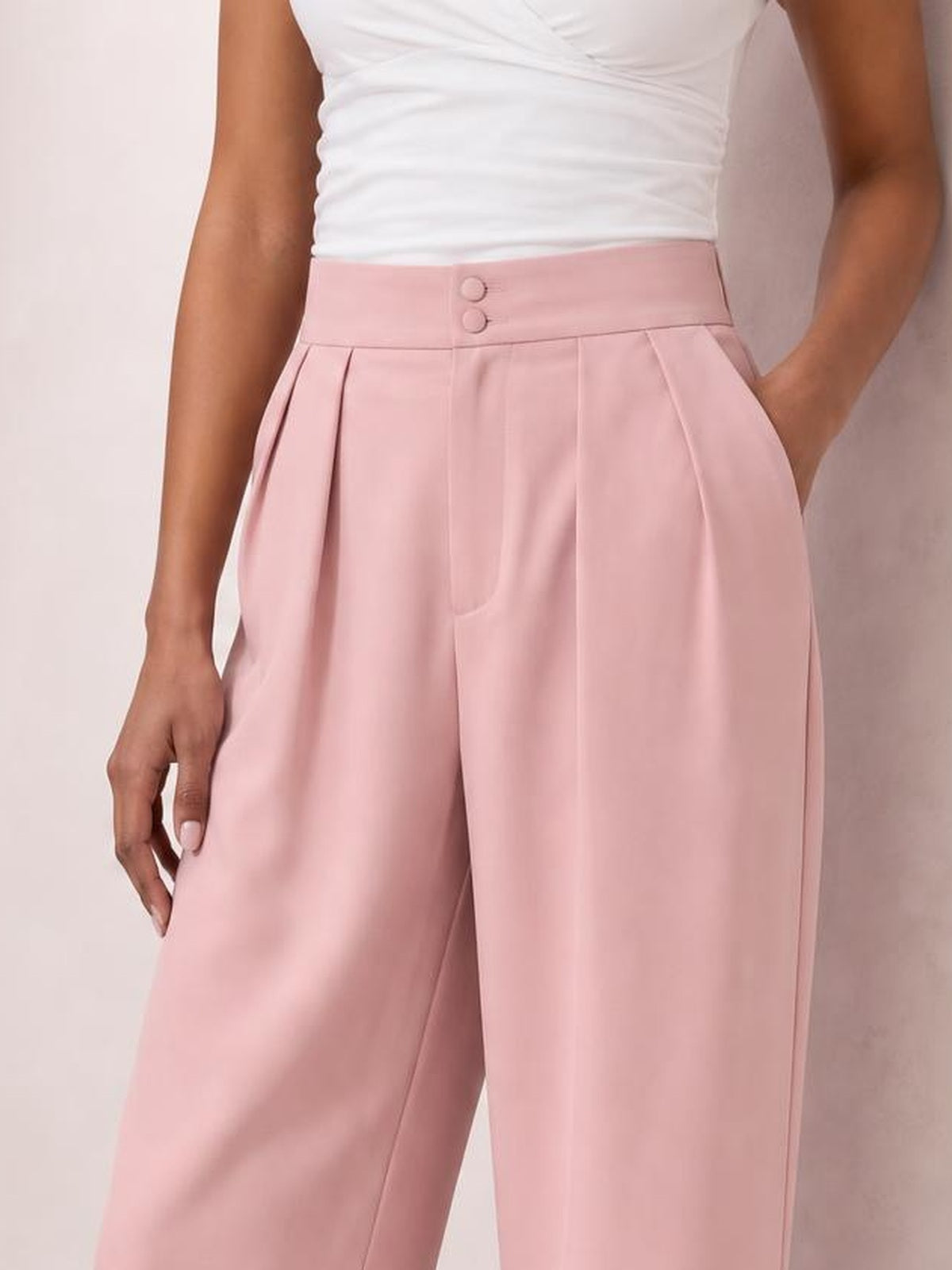 Model wears pink high-waisted pleated wide-leg pants with a buttoned waistband and side pockets, standing with one hand in a pocket.