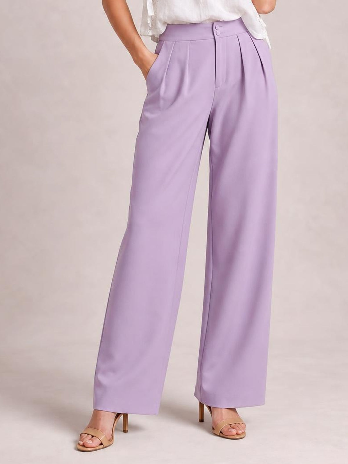 Model wearing lavender high-waisted pleated wide-leg pants with a buttoned waistband, side pockets, and a relaxed full-length silhouette.