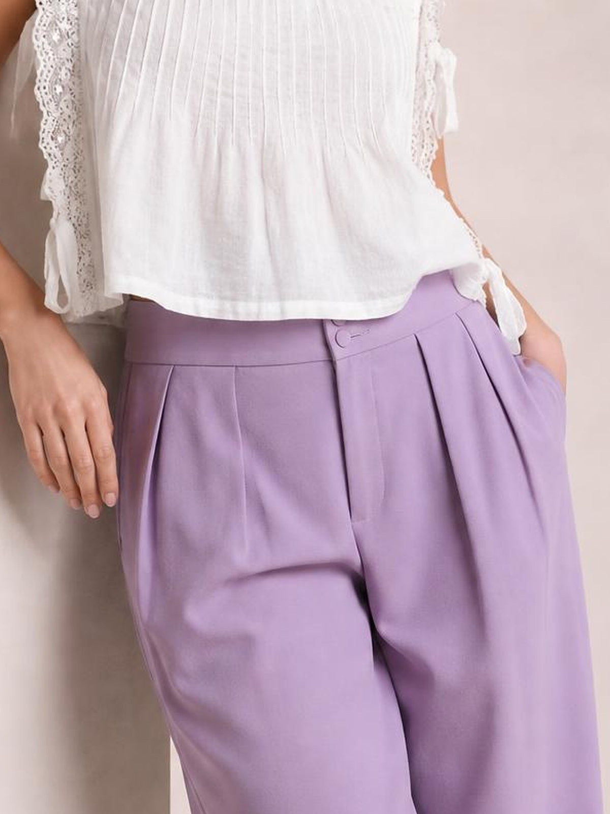 Model wearing high-waisted lavender pleated wide-leg pants with a button closure, structured waistband, and side pockets.