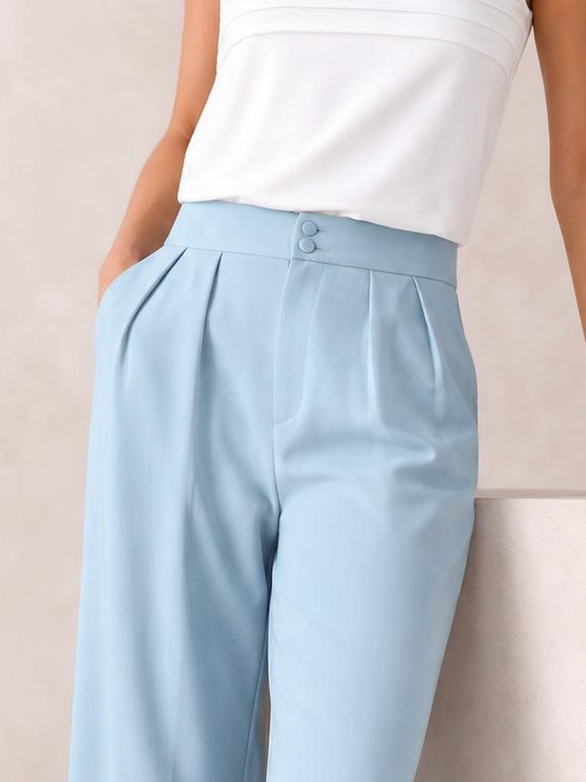 Model wears light blue high-waisted pleated wide-leg pants with a buttoned waistband and side pockets, shown with one hand in a pocket.