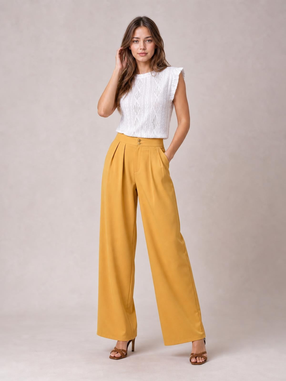 Model poses in mustard yellow high-waisted pleated wide-leg pants with a buttoned waistband, front pockets, and flowing full-length silhouette.