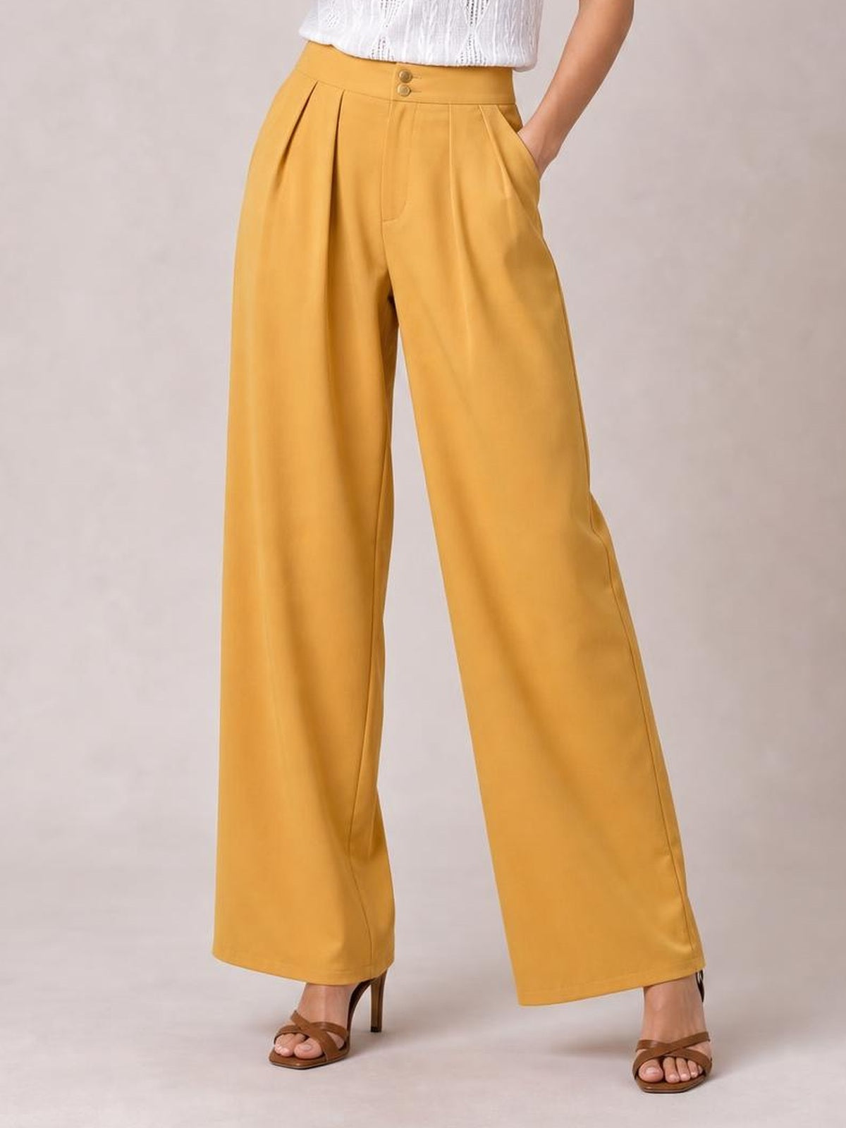 Model wearing mustard yellow high-waisted pleated wide-leg pants with button closure and side pockets, posed with one hand in a pocket.