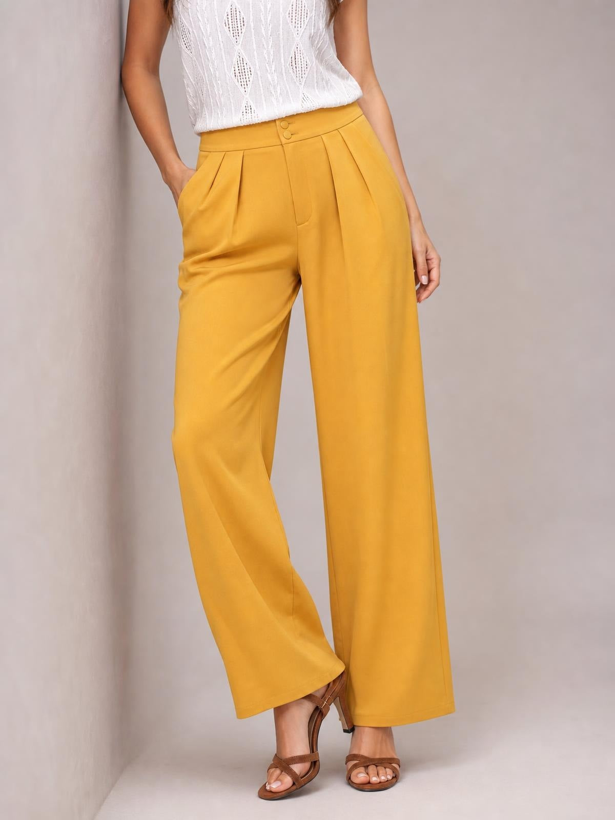Model wearing mustard yellow high-waisted pleated wide-leg pants with buttoned waistband and side pockets, posing with one hand tucked into a pocket.