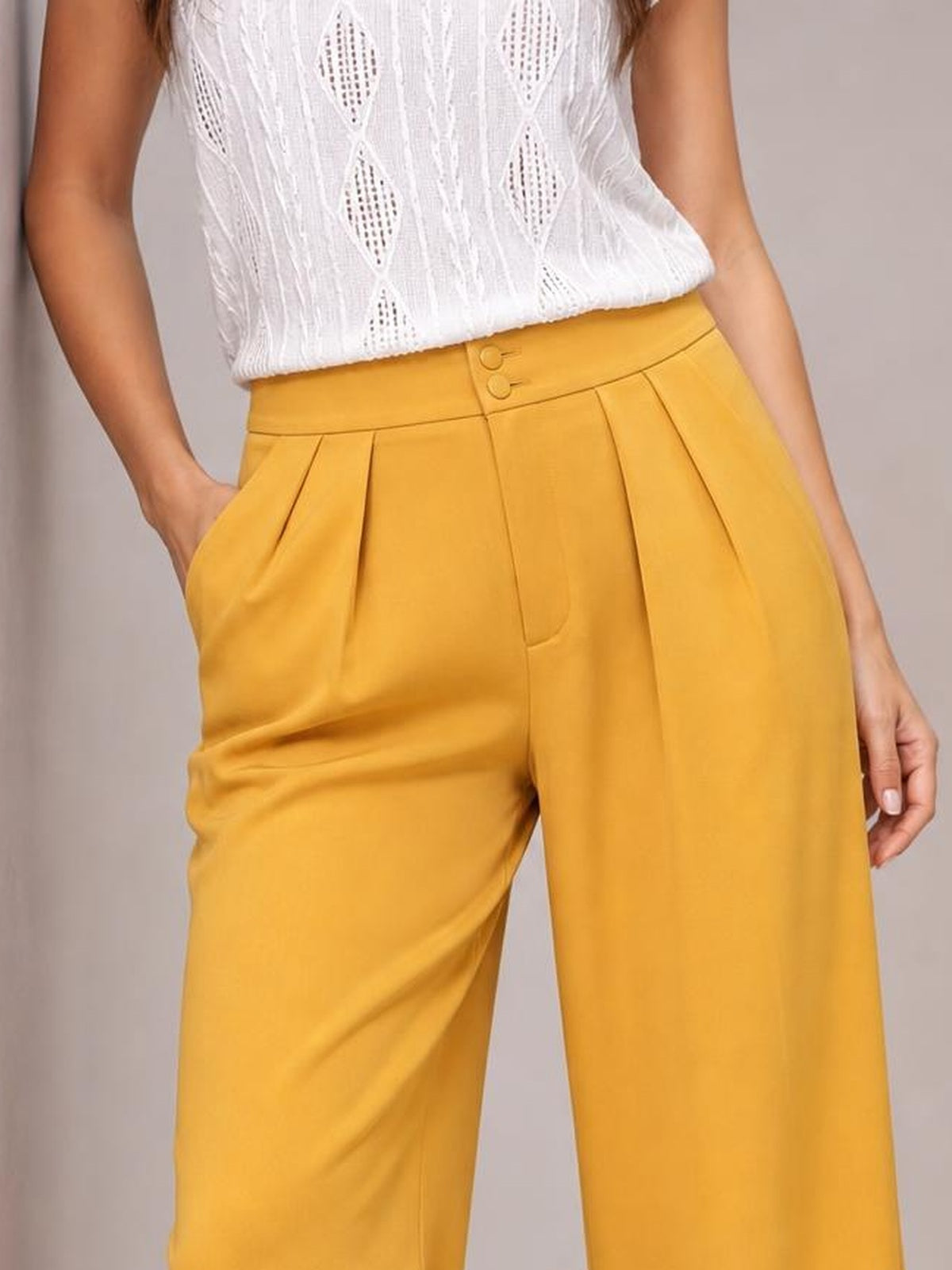A model wears mustard-yellow high-waisted pleated wide-leg pants with a buttoned waistband and side pockets, posing with one hand tucked into a pocket.