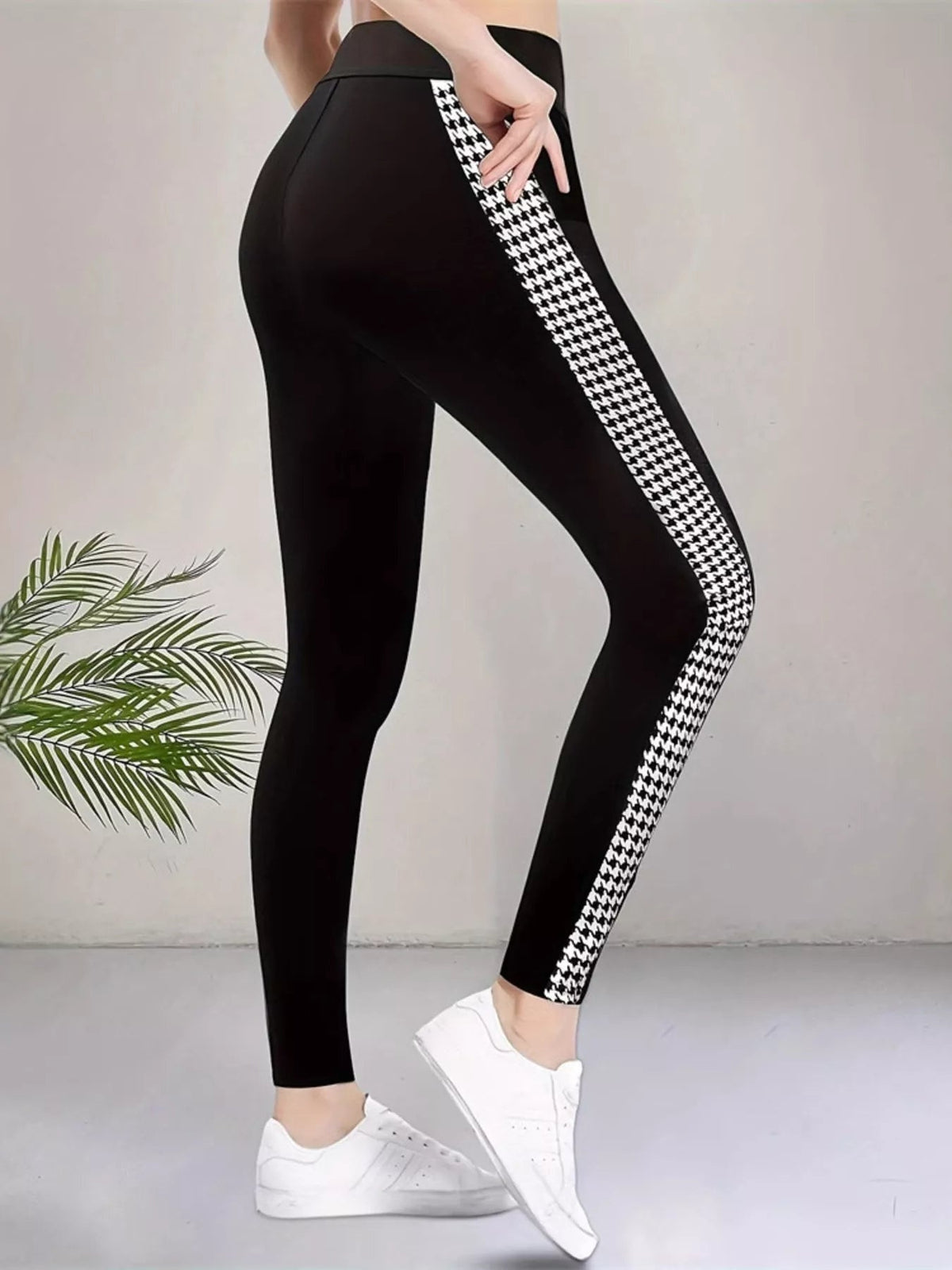 A model poses in black high-waisted active leggings with a fitted ankle-length silhouette, visible seam detail, and a bold white houndstooth side panel.