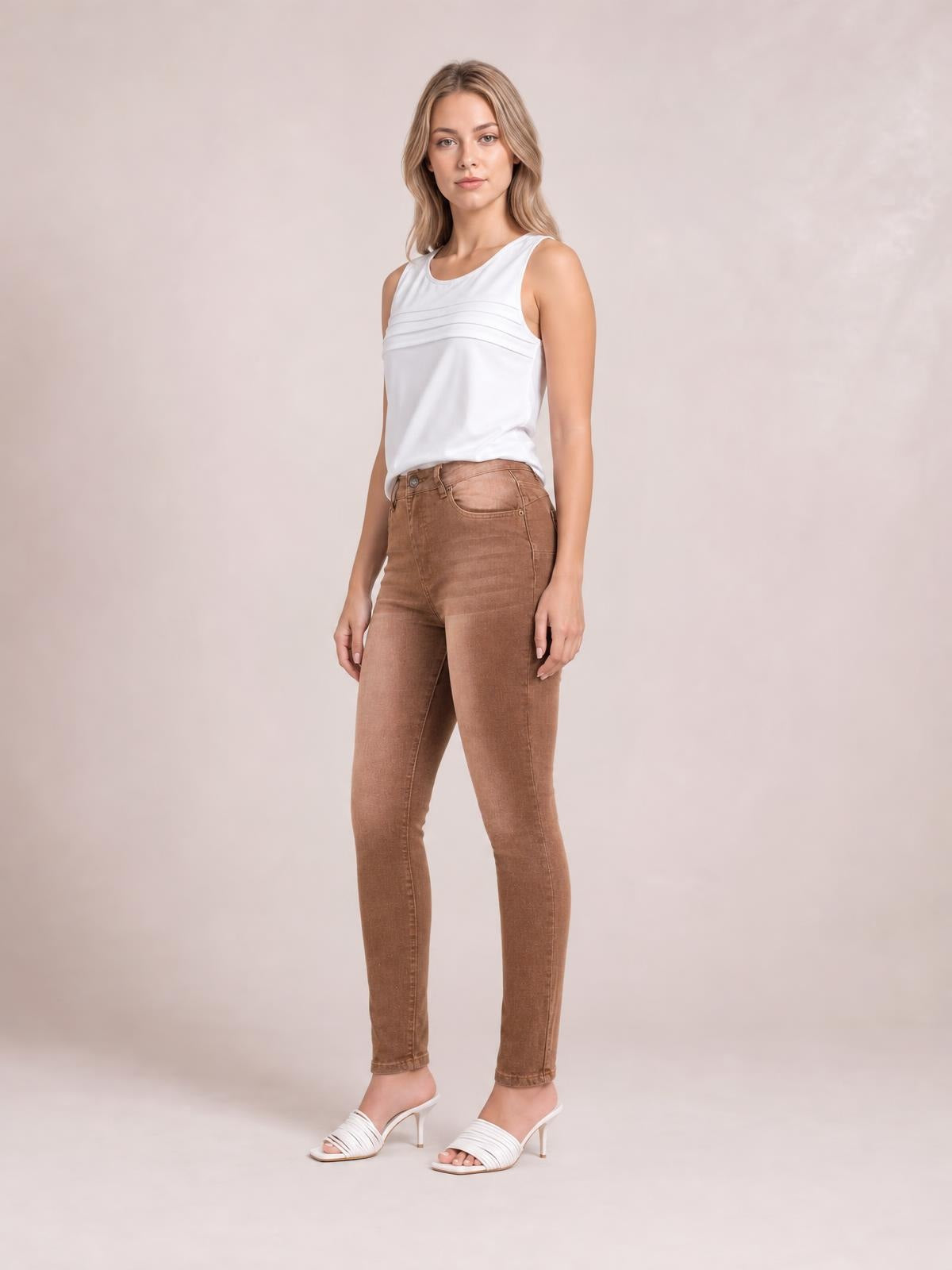 A model stands in brown high-waisted slim-fit jeans with a button closure, front pockets, fitted ankle-length legs, and a structured five-pocket construction.