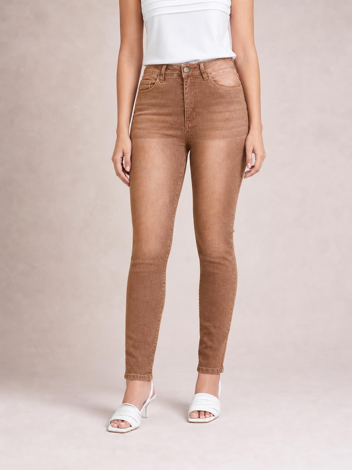 A model stands in high-waisted slim-fit jeans in warm brown, featuring a button fly, belt loops, front pockets, and a fitted ankle-length silhouette.