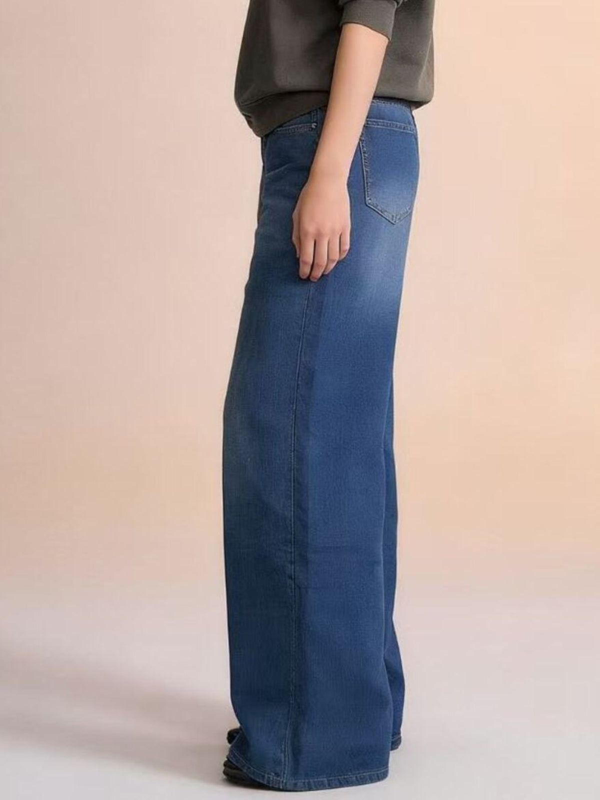 A model stands in side profile wearing blue high-waisted wide leg jeans with a relaxed silhouette, visible back pocket, belt loops, and full-length seams.