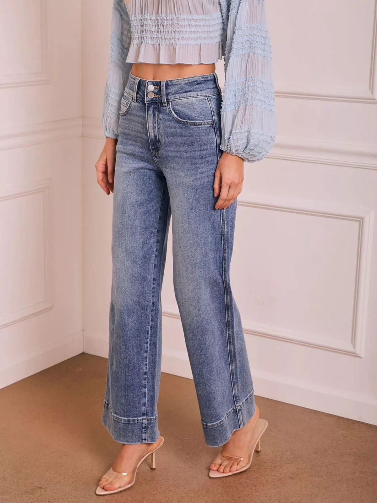 A model stands with one leg angled in light blue washed high-waisted wide-leg jeans featuring a button fly, front pockets, cat’s whisker fading, and structured hems.