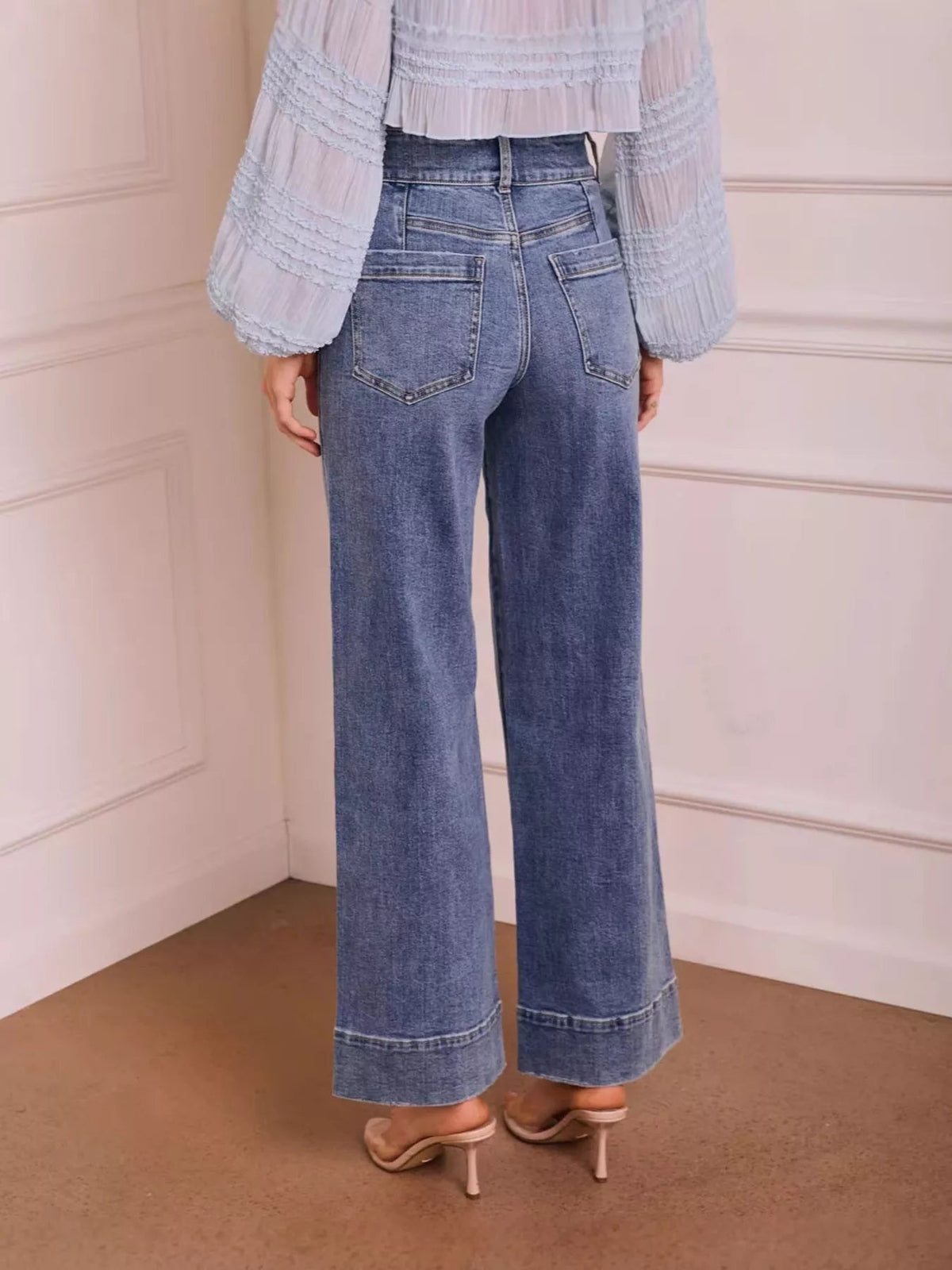 A model stands in a rear view wearing high-waisted wide-leg jeans in washed blue denim with two back patch pockets, contrast stitching, and wide seamed hems.