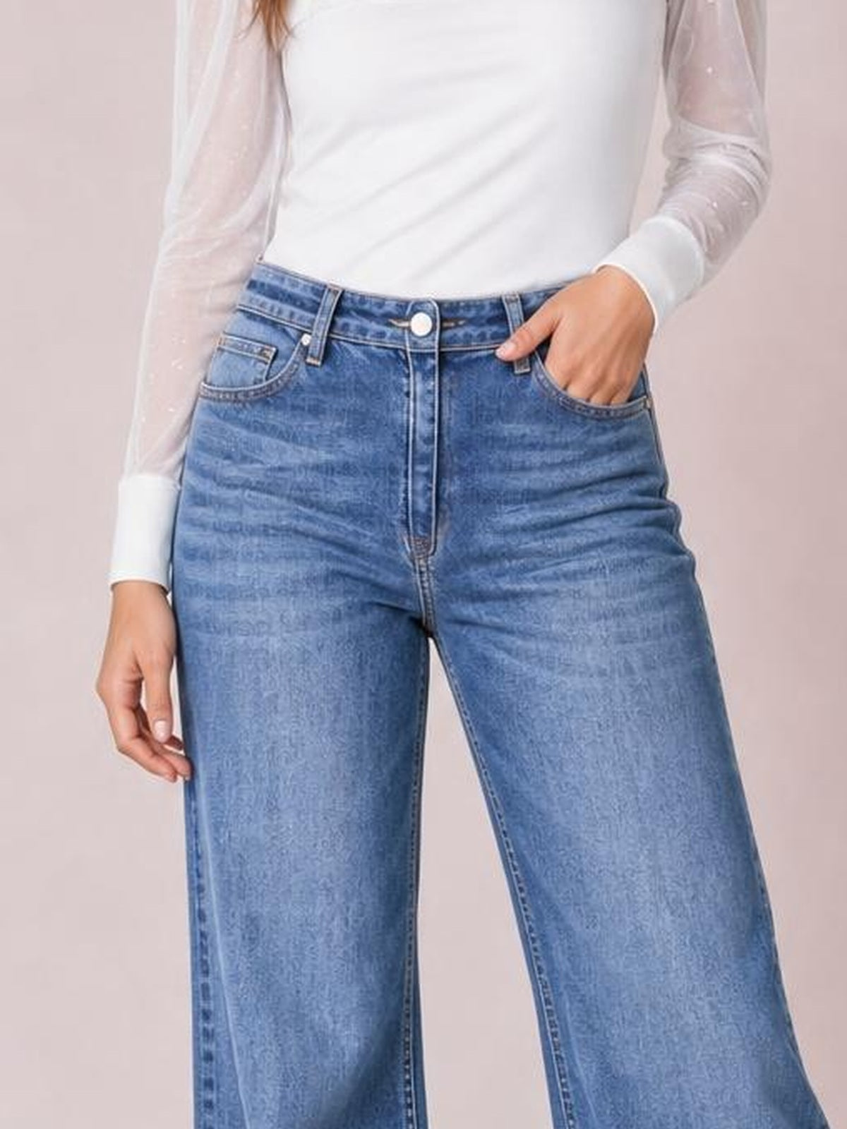 Model poses with one hand in the pocket of light blue washed high-waisted wide-leg jeans, showing belt loops, front pockets, button closure, and zip fly.