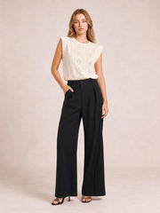 A model poses in black high-waisted wide-leg pants with a front button closure, pleated waist, relaxed silhouette, and floor-skimming hems.