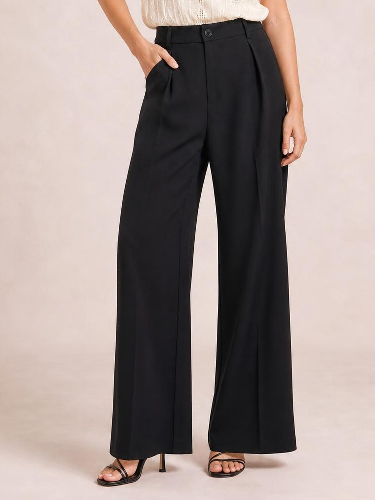 A model poses in black high-waisted wide-leg pants with a buttoned waistband, front pleats, side pockets, and a fluid full-length silhouette.