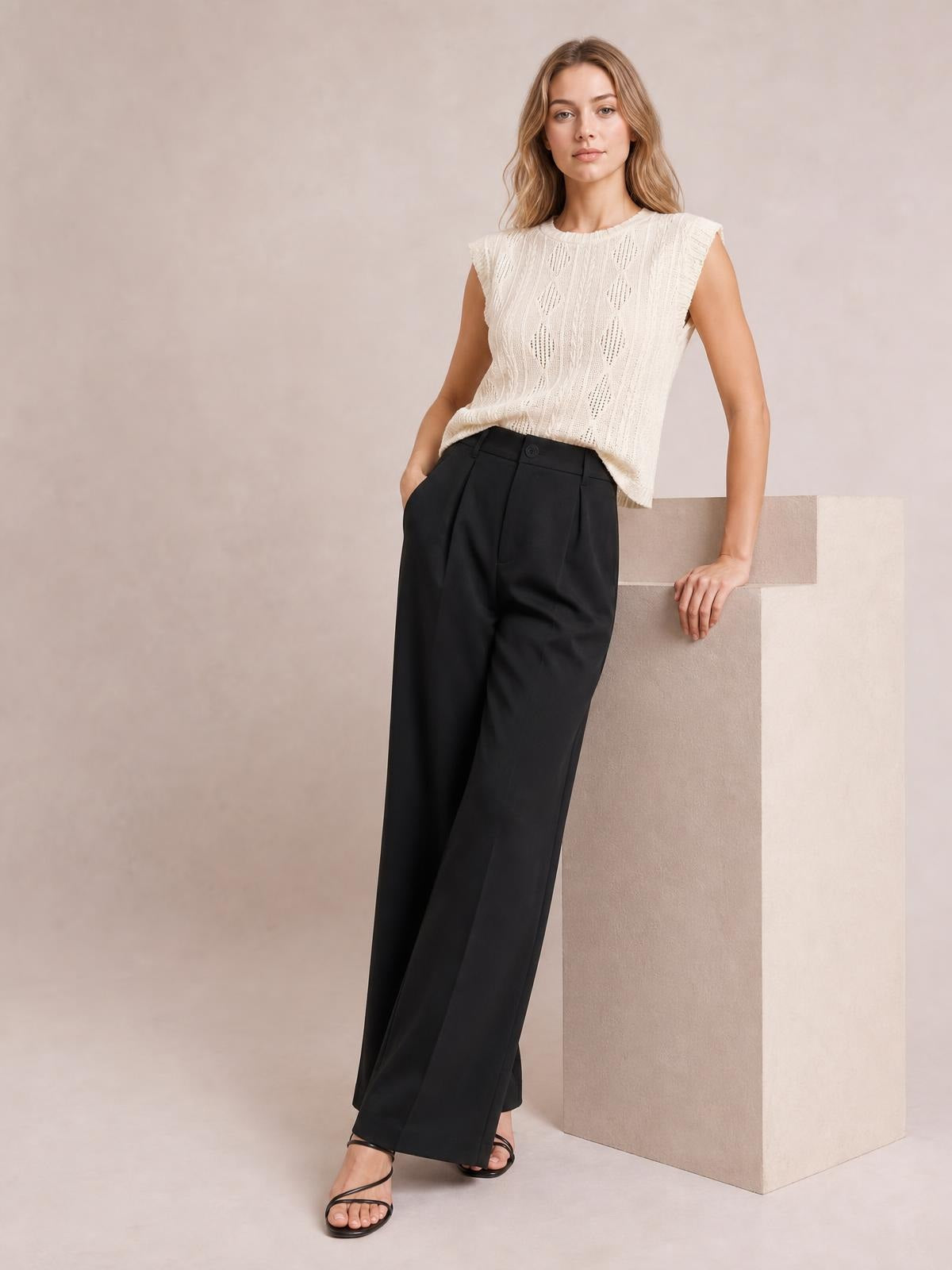 Model wearing black high-waisted wide-leg pants with front pleats and a button closure, posed with one hand in a pocket.