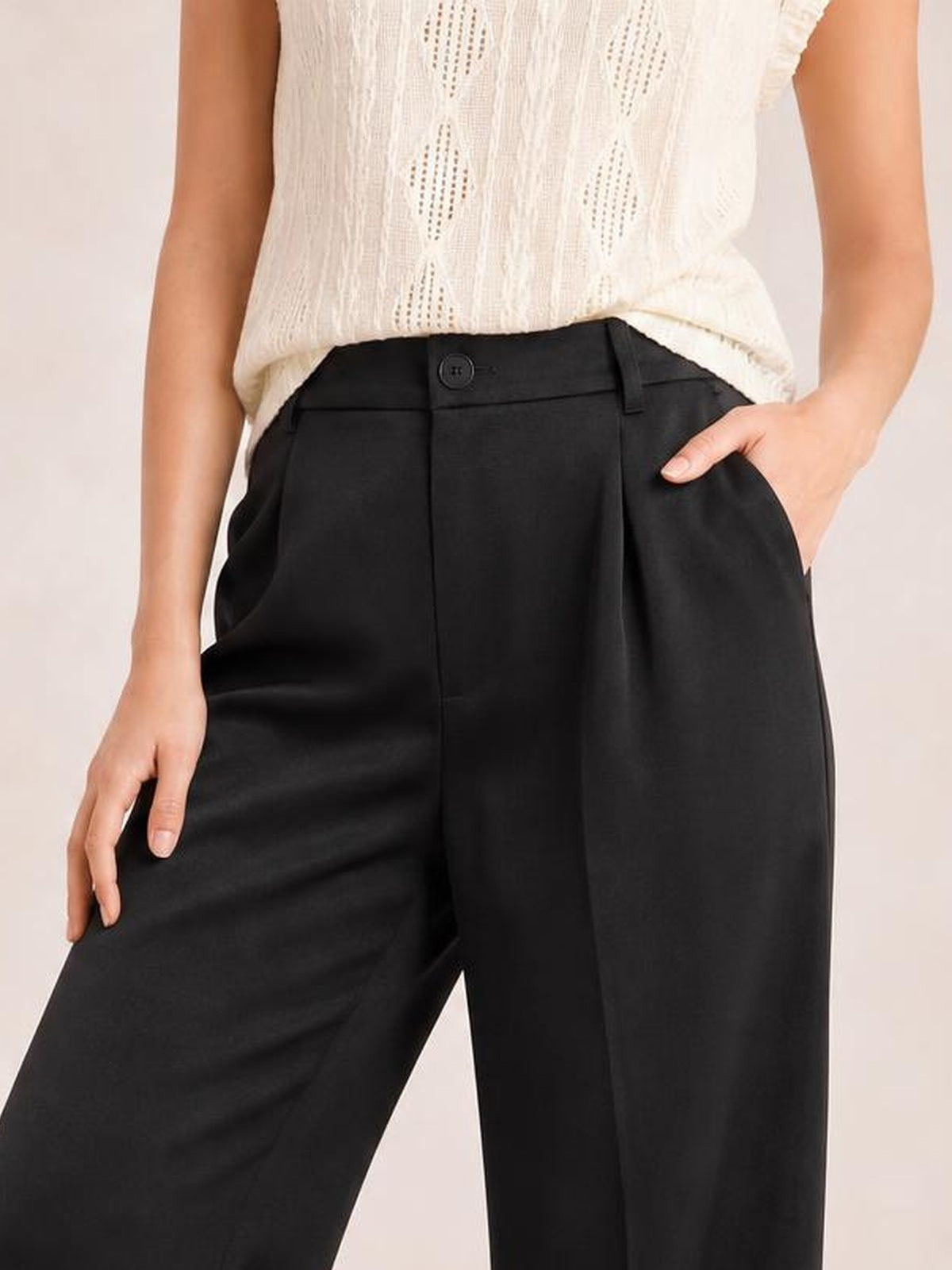 Model wears black high-waisted wide-leg pants with a button closure, belt loops, structured waistband, and relaxed silhouette, posing with one hand in a pocket.