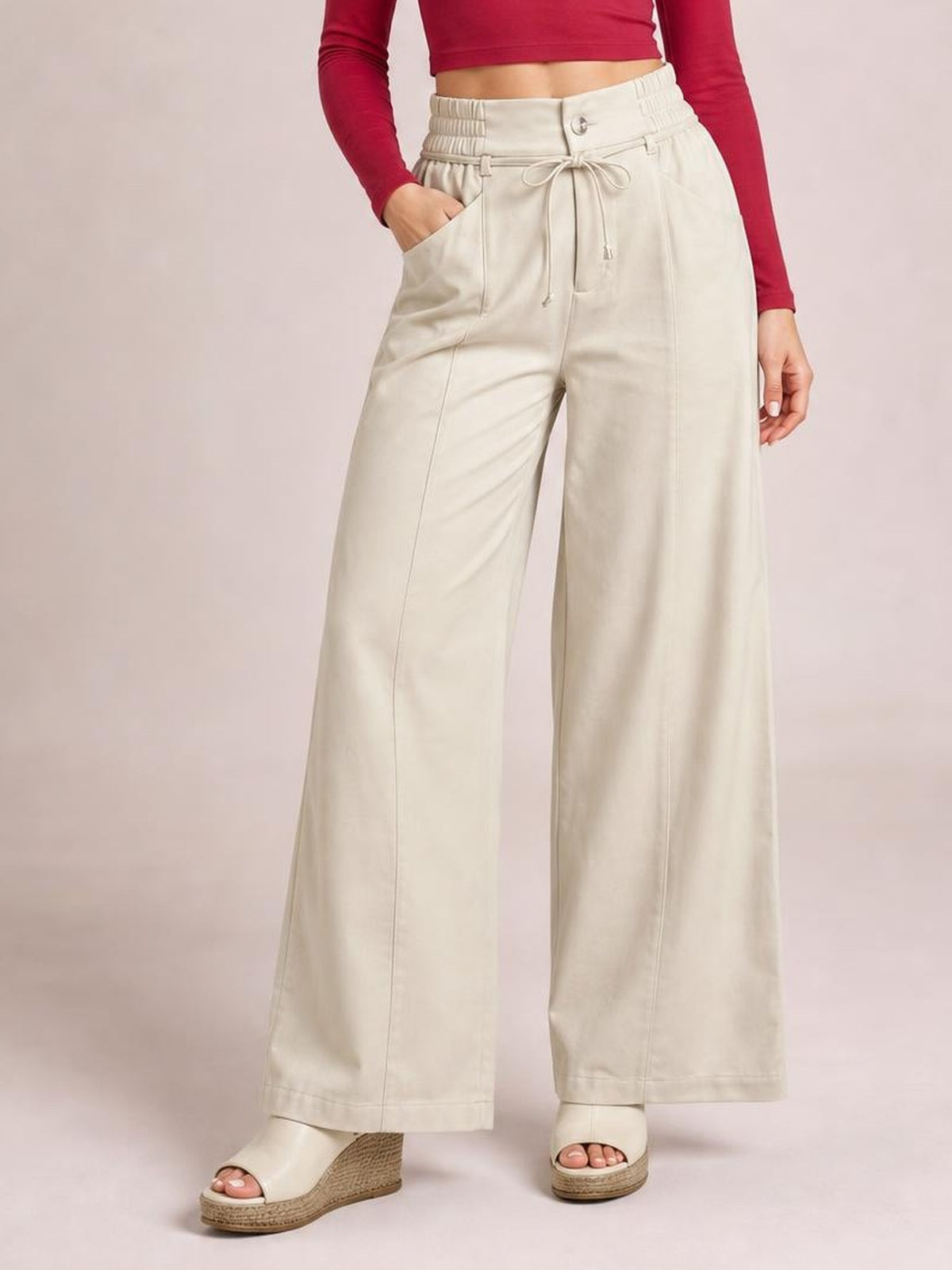 Model poses with one hand in a pocket, wearing cream high-waisted wide-leg pants with a buttoned waist, tied drawstring, and full-length silhouette.
