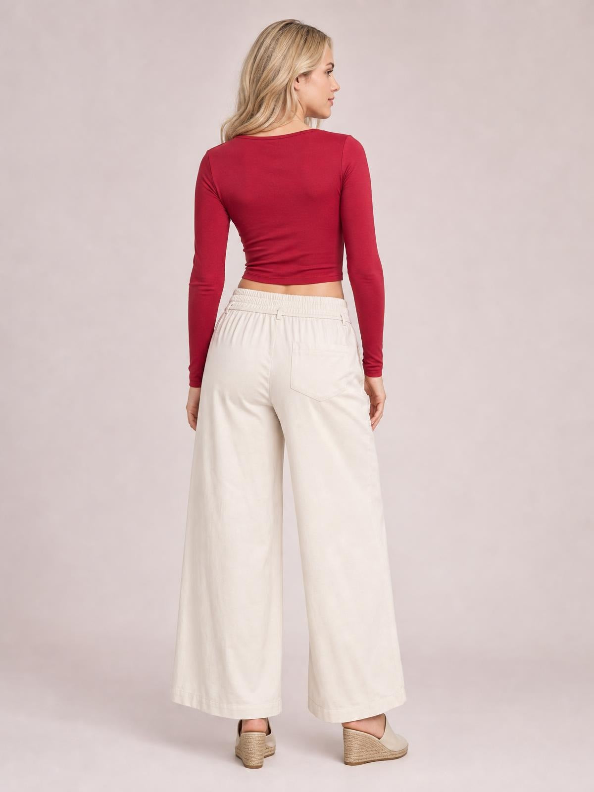 A model turns to show cream high-waisted wide-leg pants with a gathered waistband, relaxed silhouette, and visible stitched back pockets.